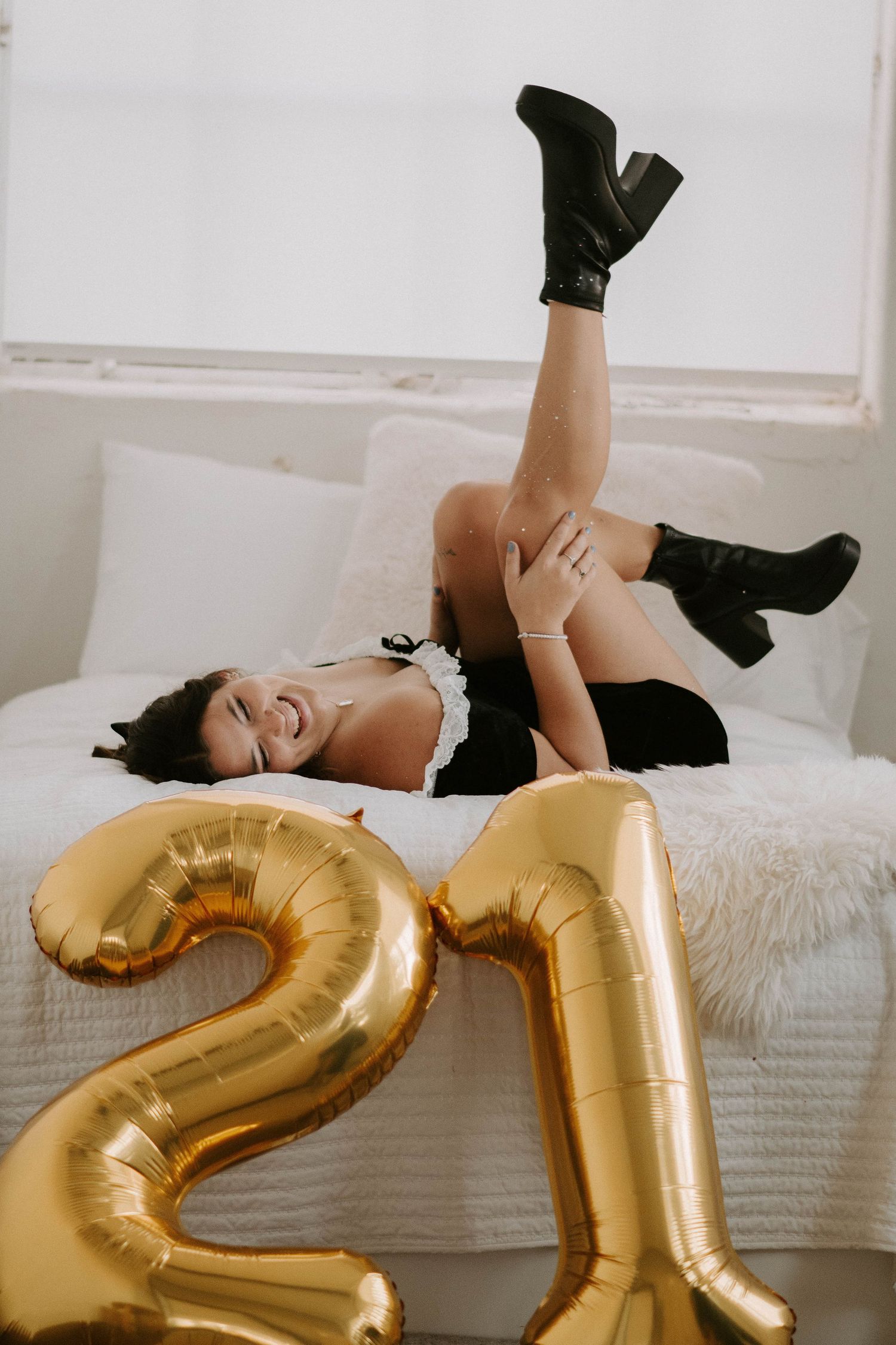 Birthday celebration scene with gold number 21 balloons and black boots on a white bed.