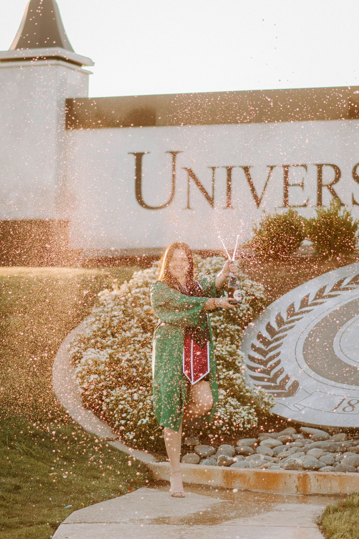 Graduate celebrating near university sign with water splashing in golden sunlight.