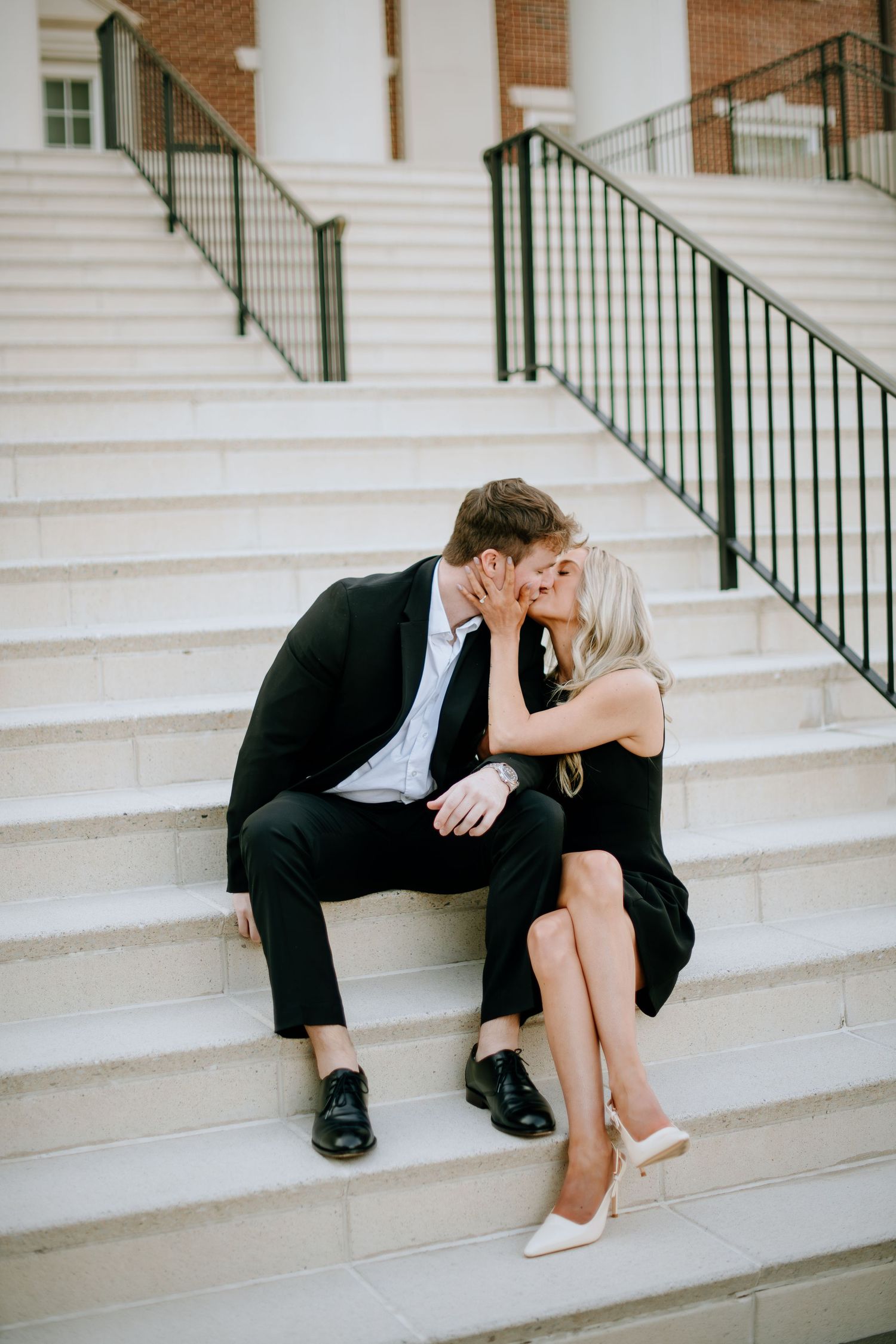 A couple shares an intimate moment while sitting on white stone steps with black railings outside a brick building.