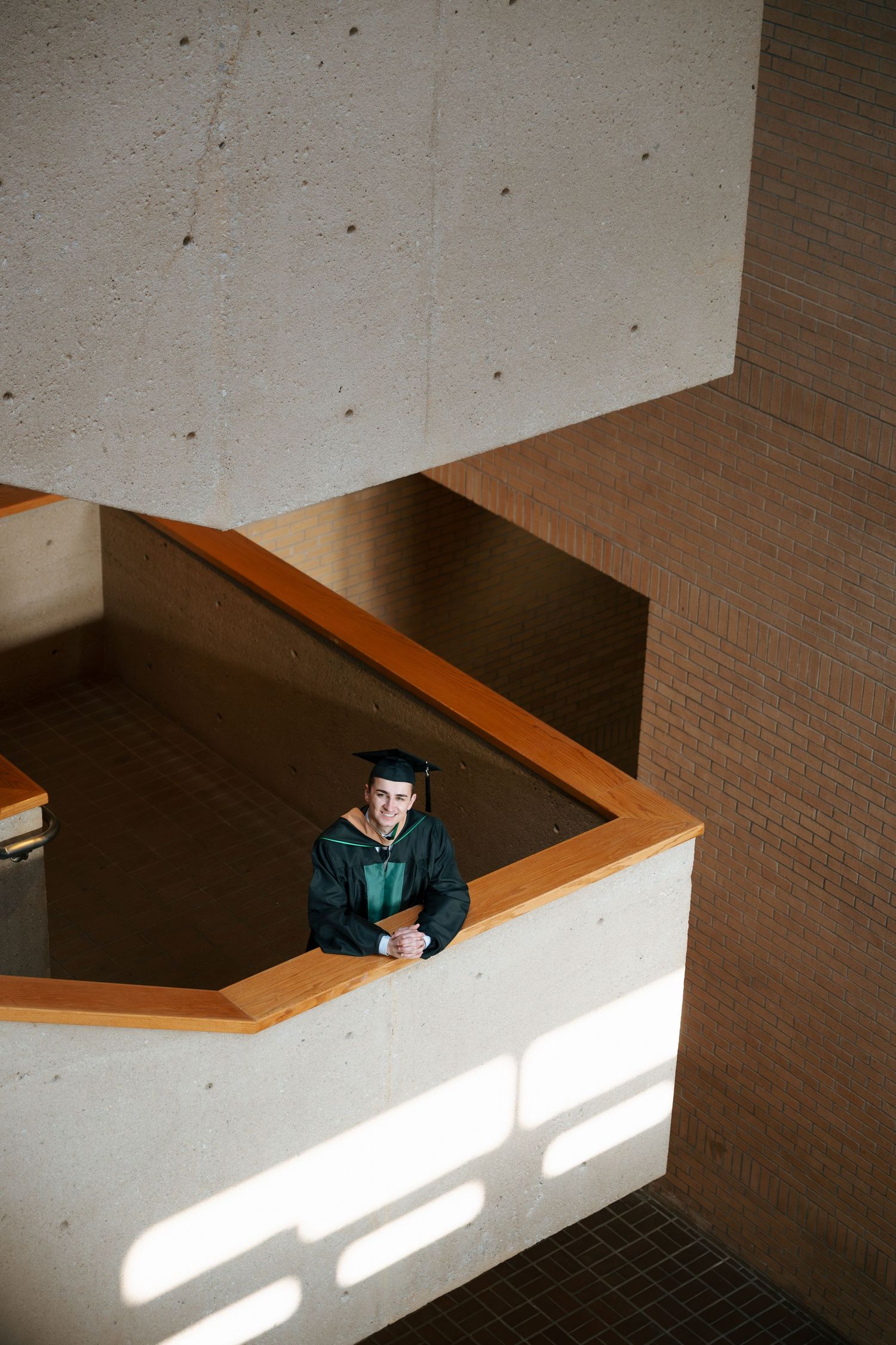 Graduate in green cap and gown stands on a modern building balcony, viewed from above in dramatic architectural setting.