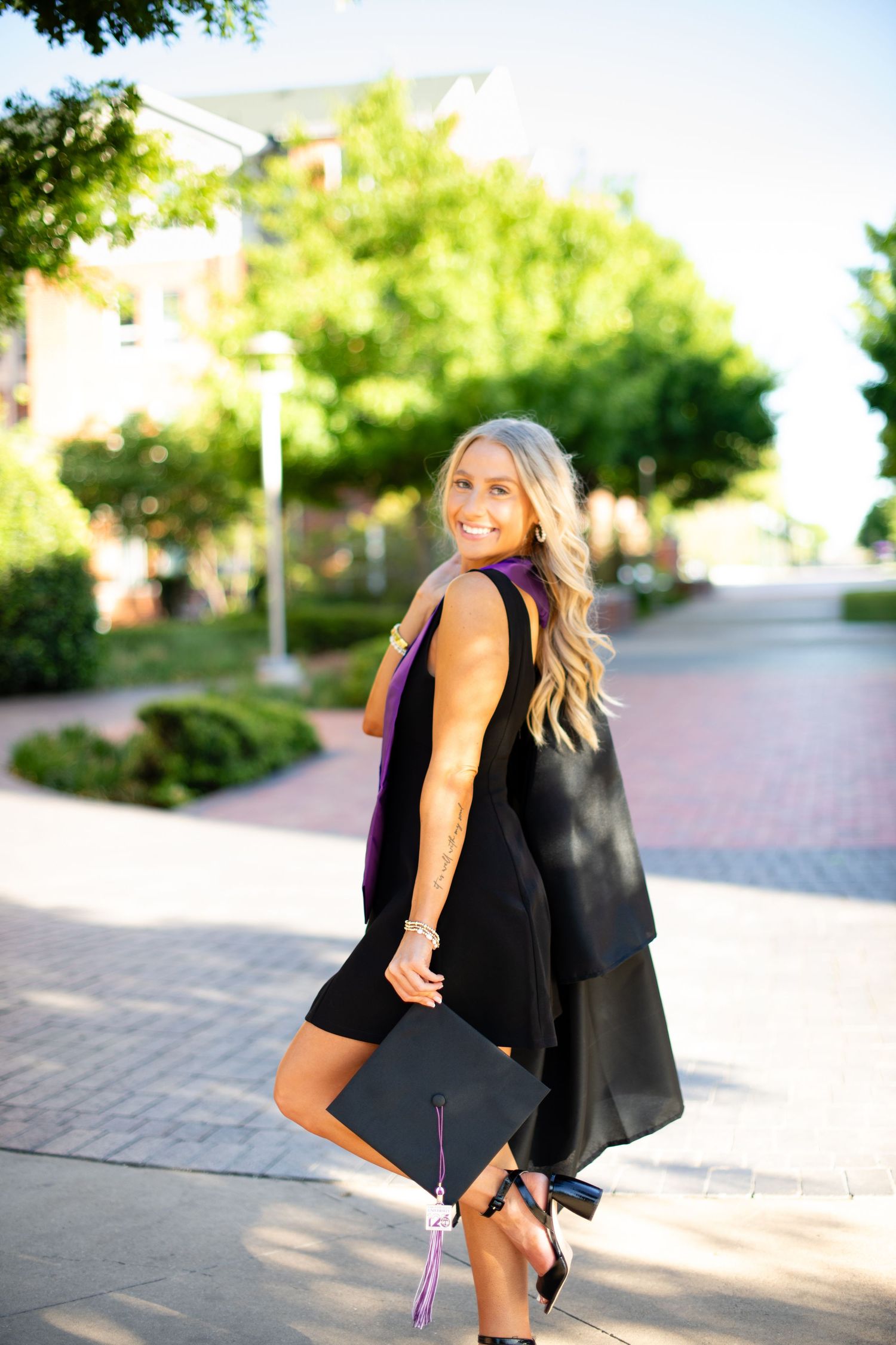 A graduation photo featuring a black cap and gown against a sunlit brick path and green trees.
