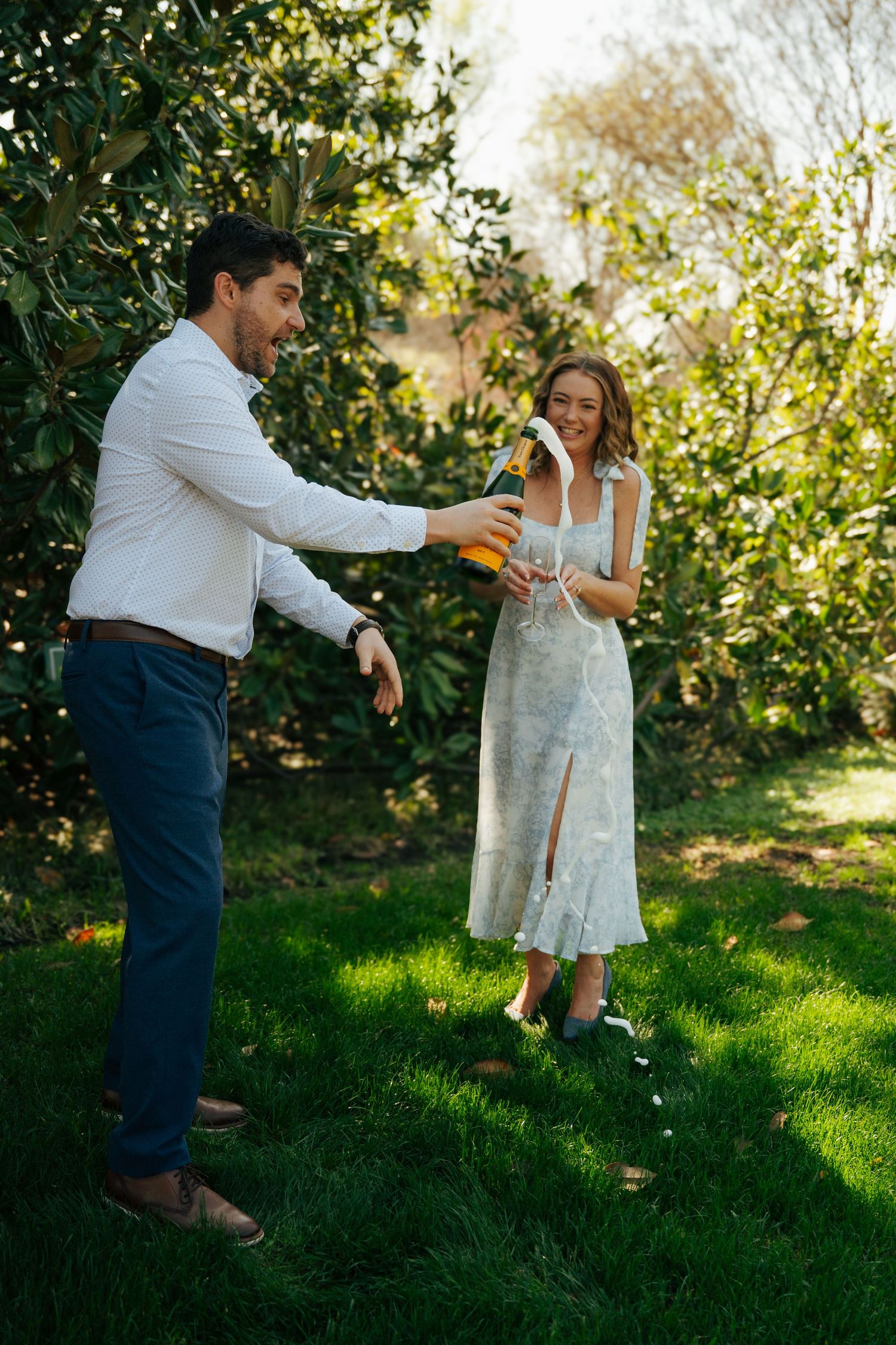 Couple celebrating outdoors on green grass, man pouring champagne for woman in floral dress near lush garden trees.
