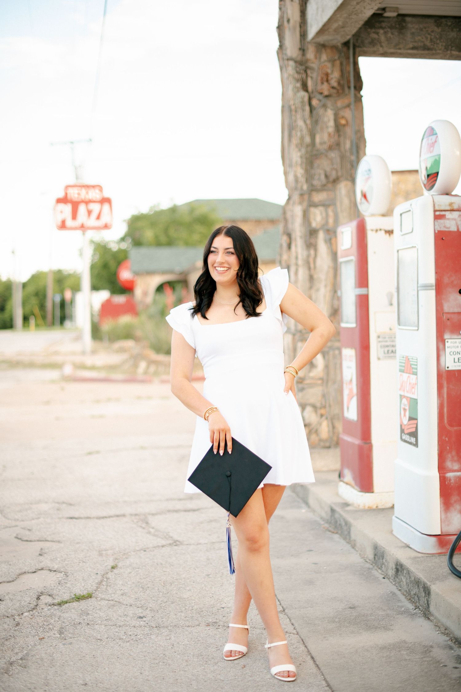 Graduate in white dress holds cap near vintage gas station.
