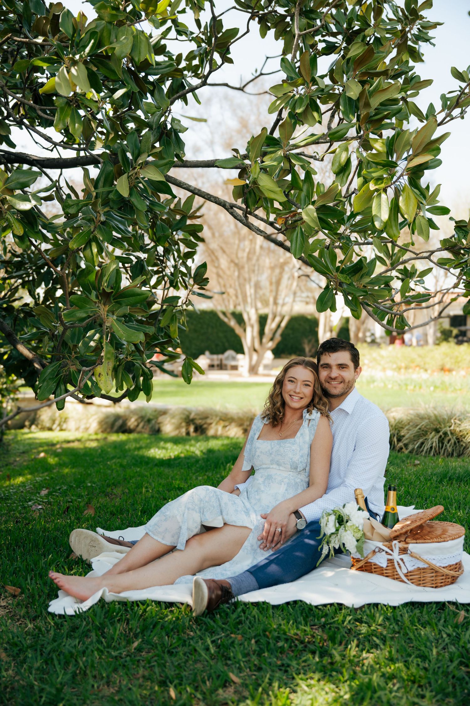Couple sitting on white blanket under magnolia tree at outdoor picnic with champagne and basket.
