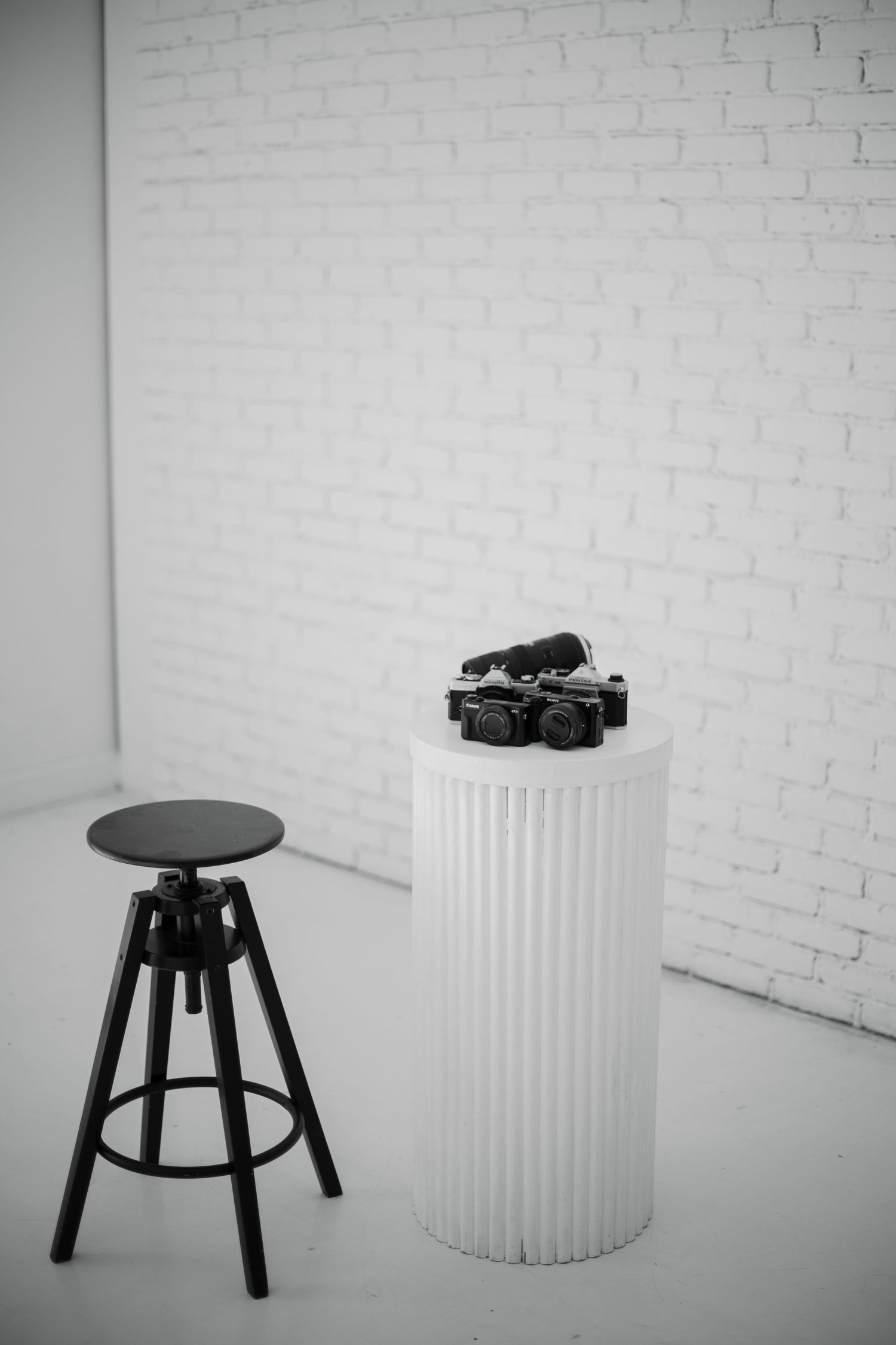 Four black and white minimalist photos showing a black stool and white cylindrical pedestal against a white brick wall.