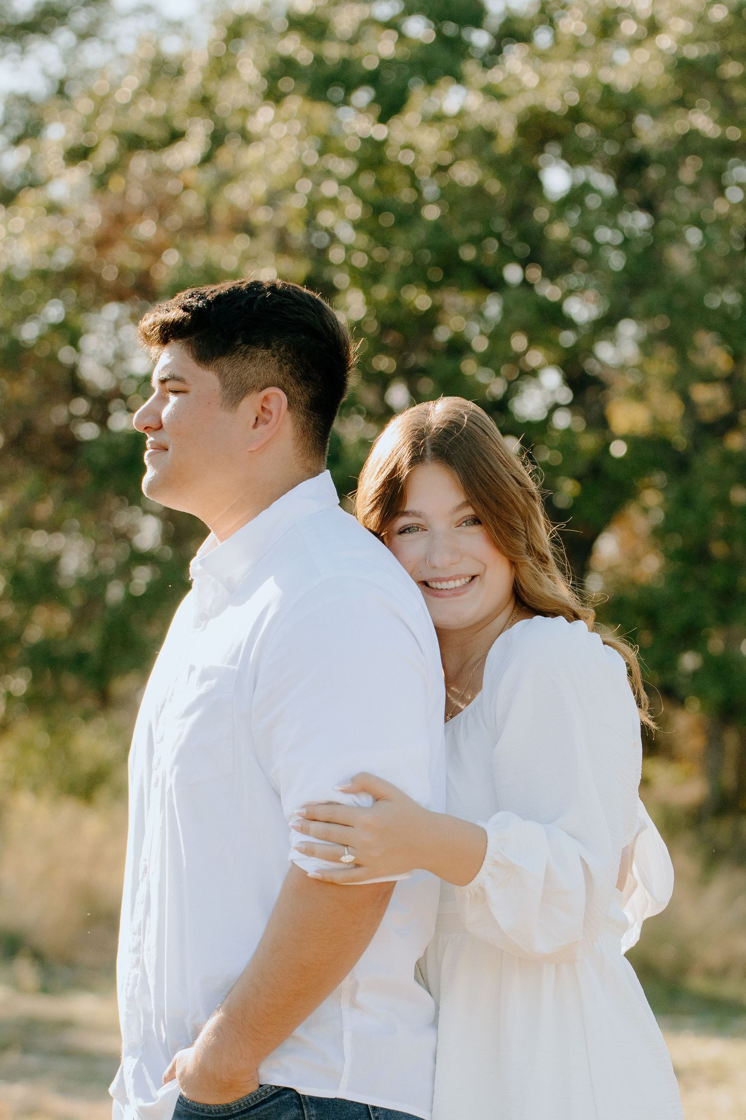 A couple in white outfits embraces outdoors on a sunny day with trees and natural lighting in the background.