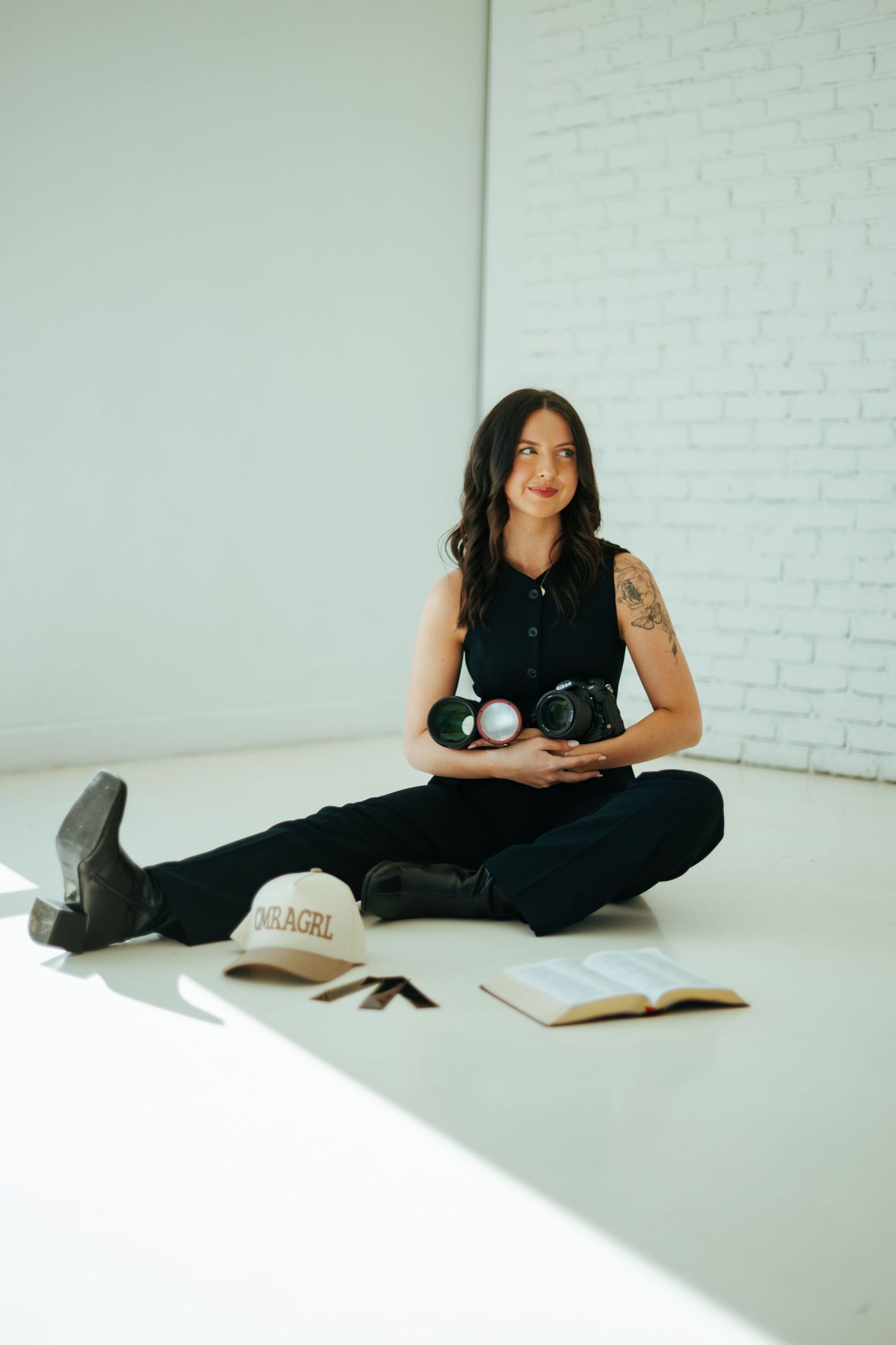 Person sitting on white floor with camera, book, and baseball cap against white brick wall in casual setting.