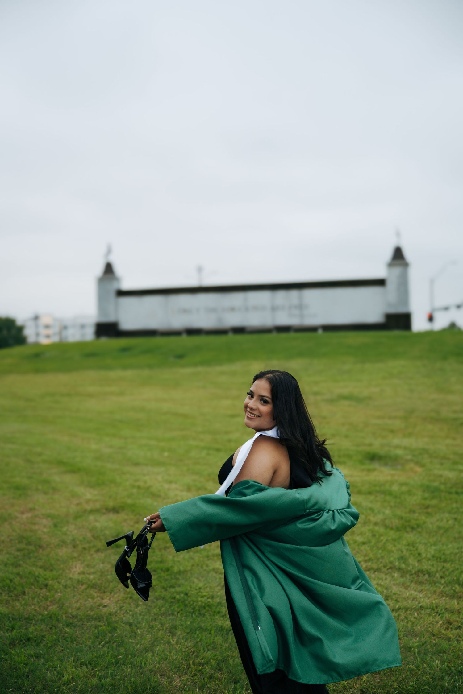 Woman in green blazer holding heels on a grassy hill with a white building in the background.