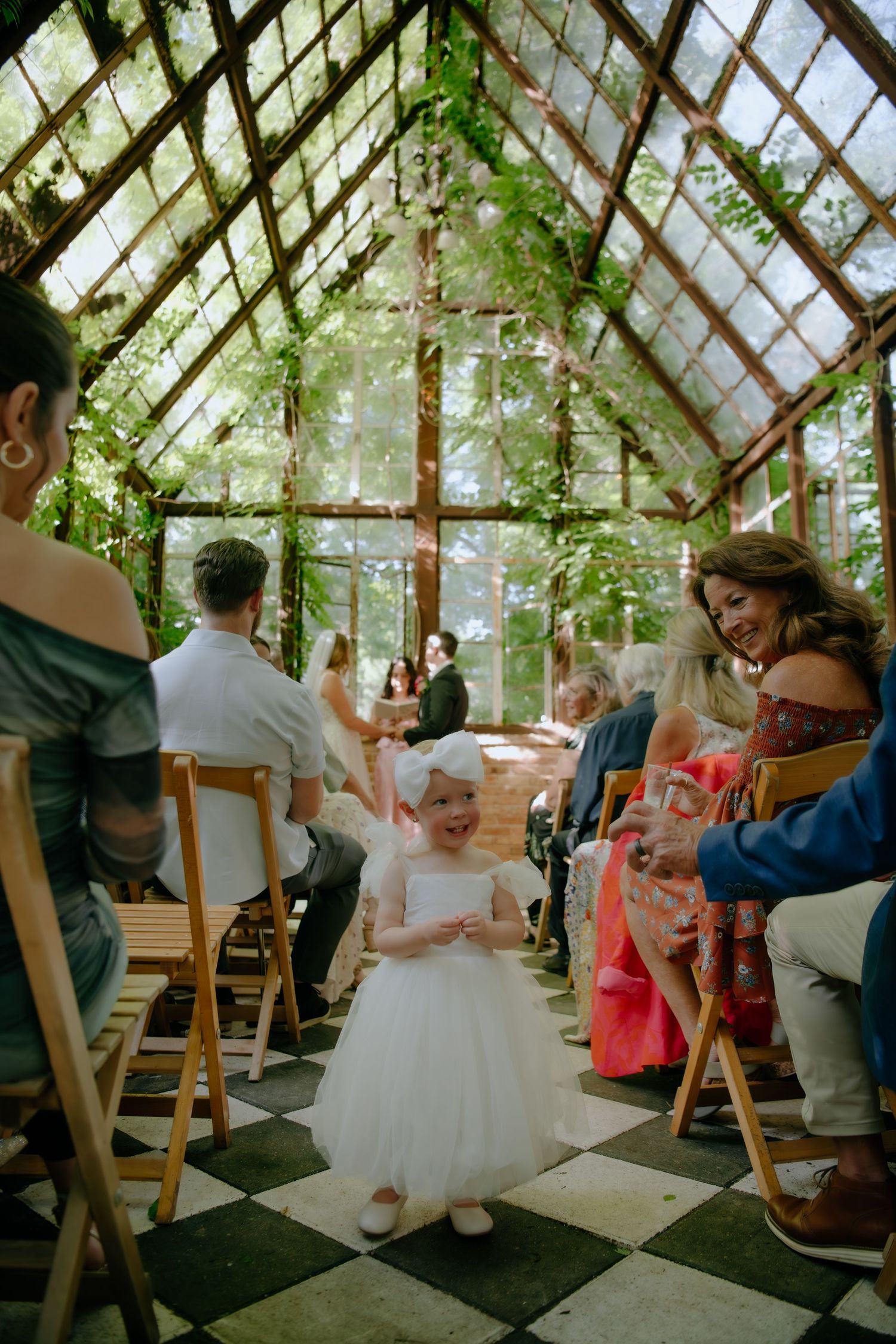 Wedding ceremony guests seated in wooden chairs inside a naturally lit greenhouse space.