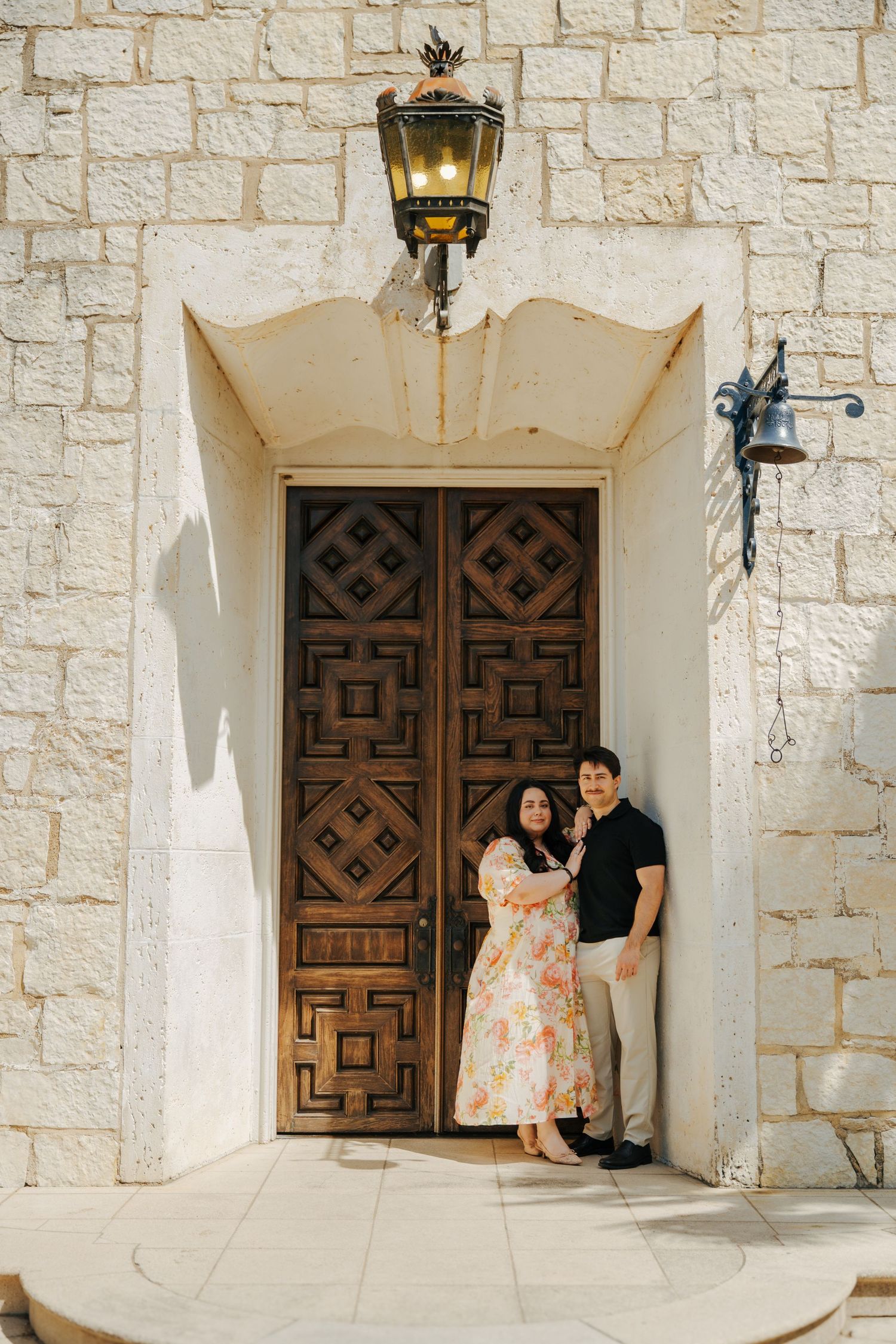 Couple posing in front of ornate wooden doors of a stone building with vintage lantern overhead.