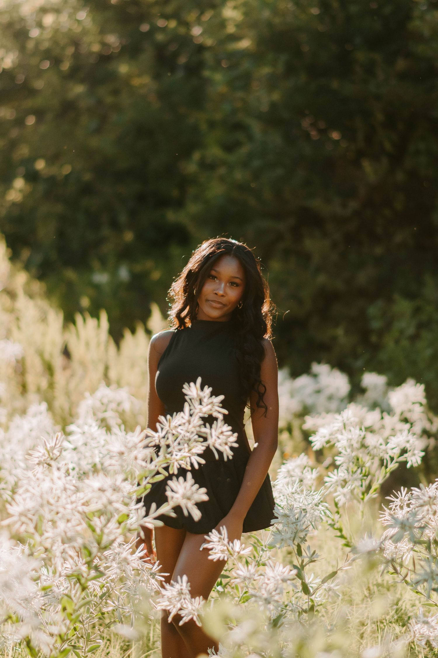 Black dress stands amid field of delicate white wildflowers at sunset.