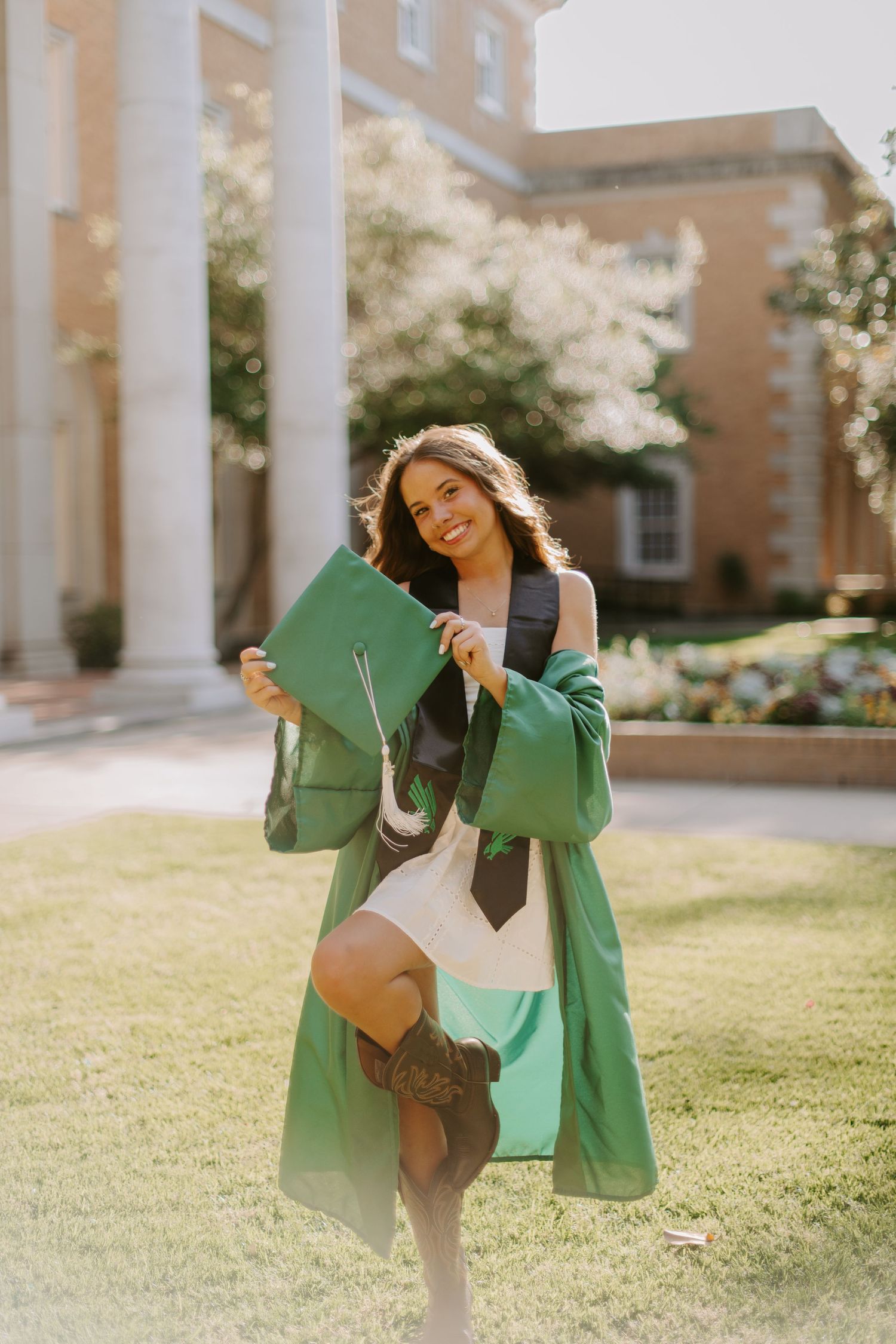 Graduate in green gown twirls joyfully in front of columned building.
