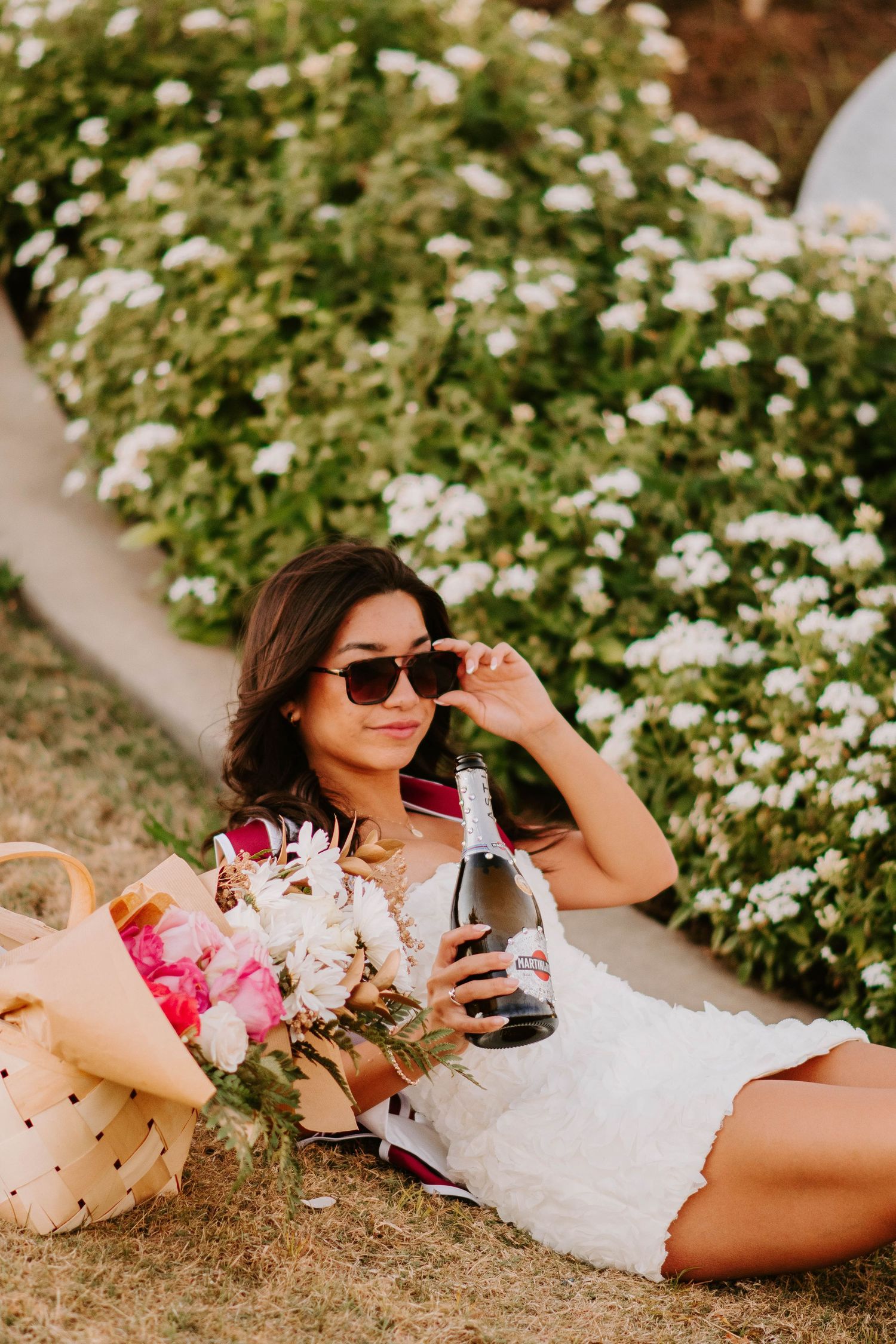 Person relaxing on grass with flowers, sunglasses, and drink on sunny day.