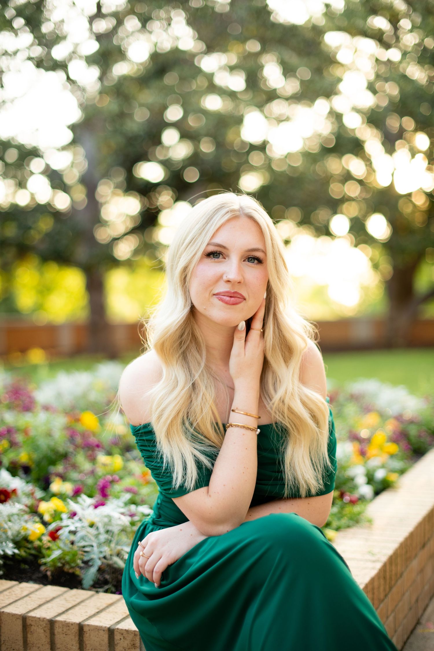 Woman in green dress sits on bench near garden with bokeh background.