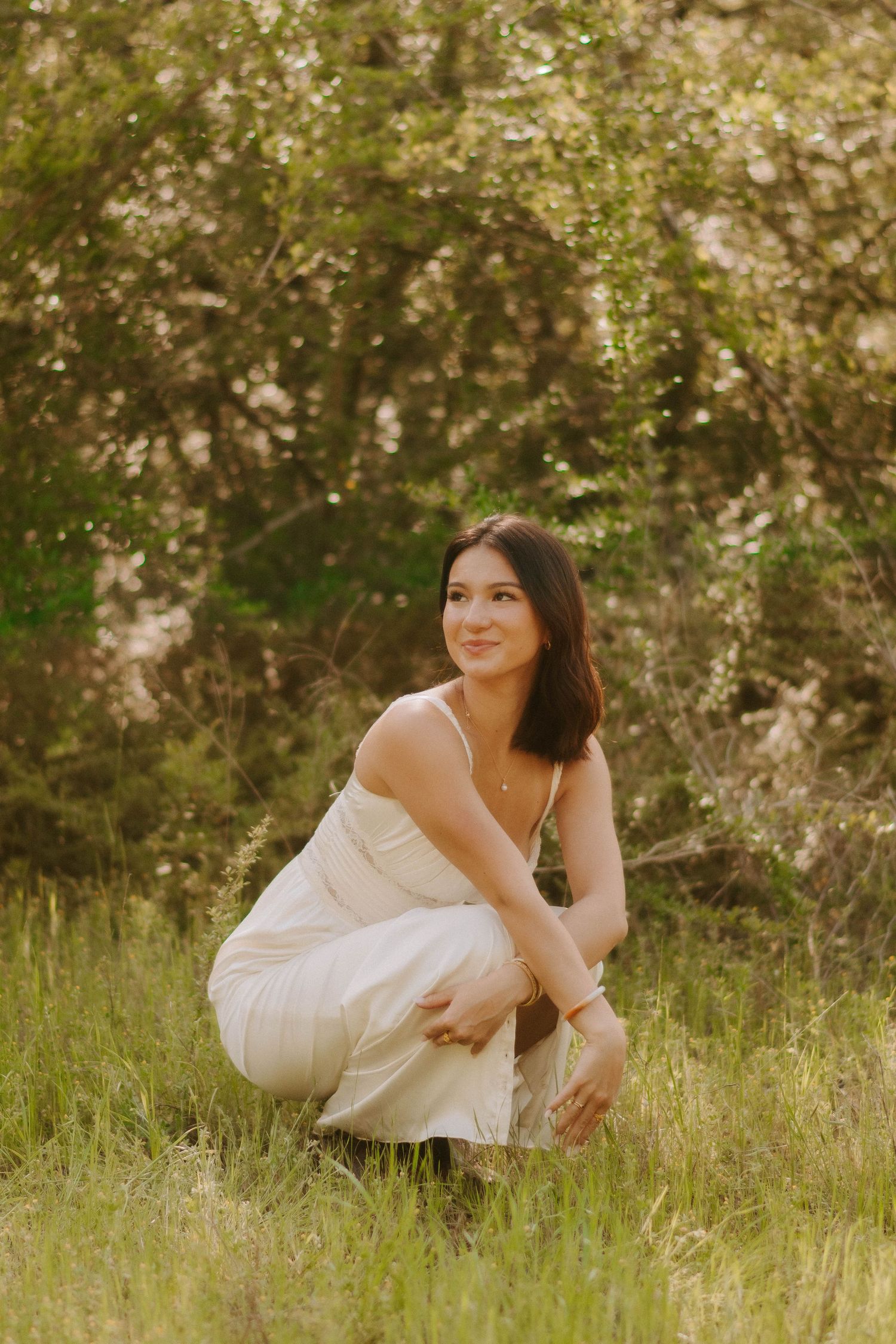 A person in a white dress crouches in a sunlit meadow with flowering trees in the background.