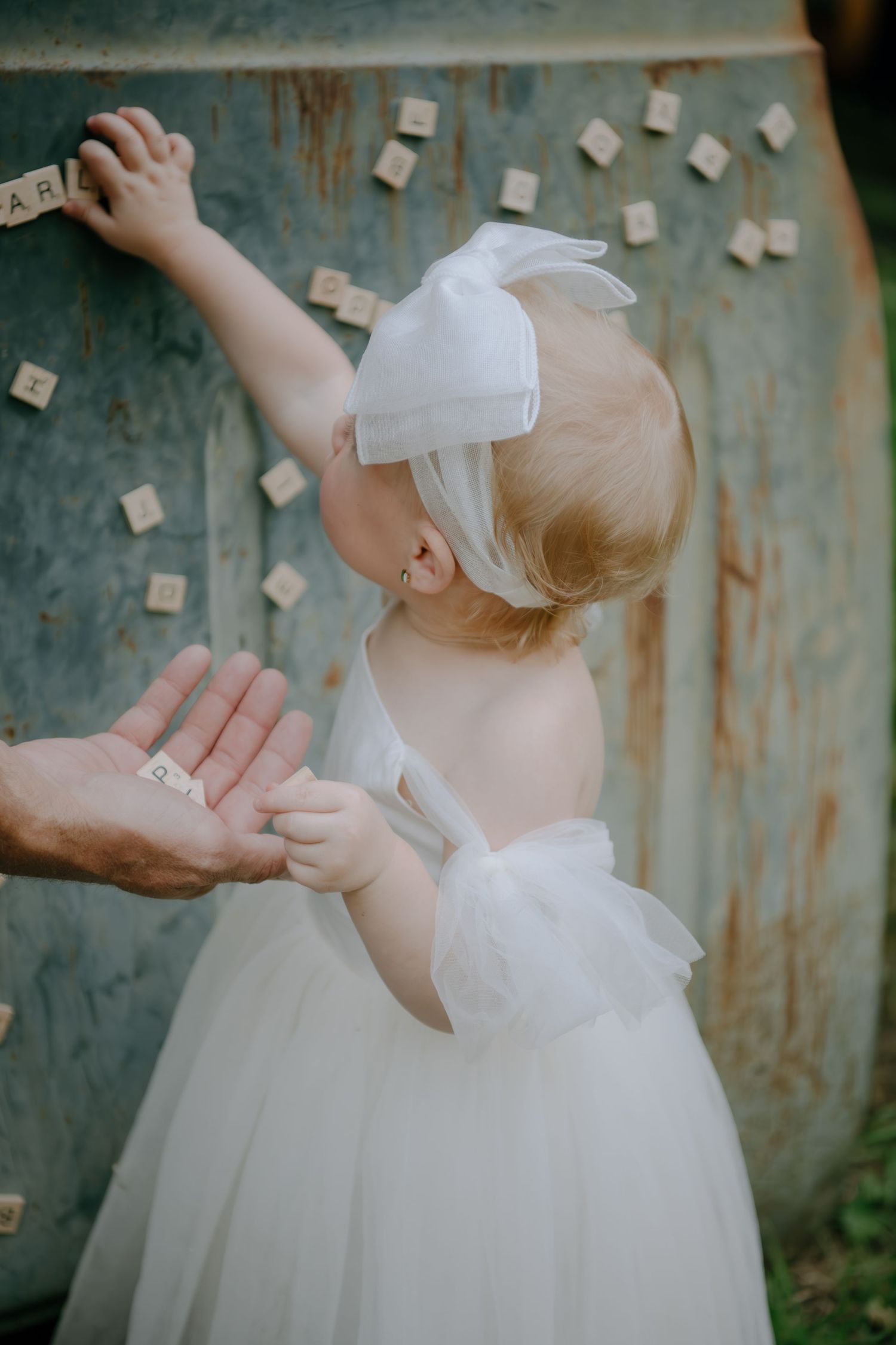 Young child in white dress and bow reaching up toward wooden letter blocks.