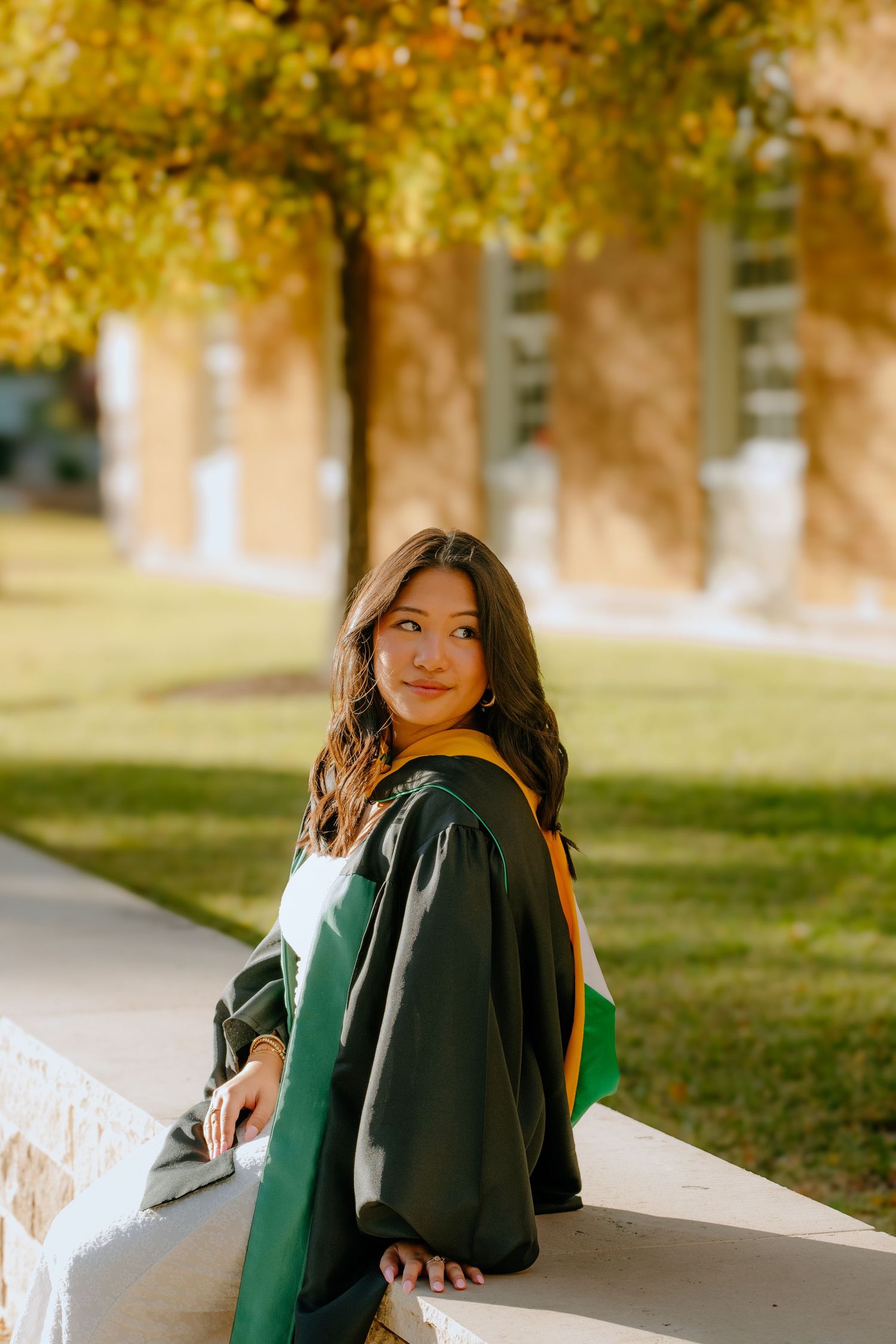 A graduate in a dark green academic gown sits on a ledge during autumn with golden trees in the background.