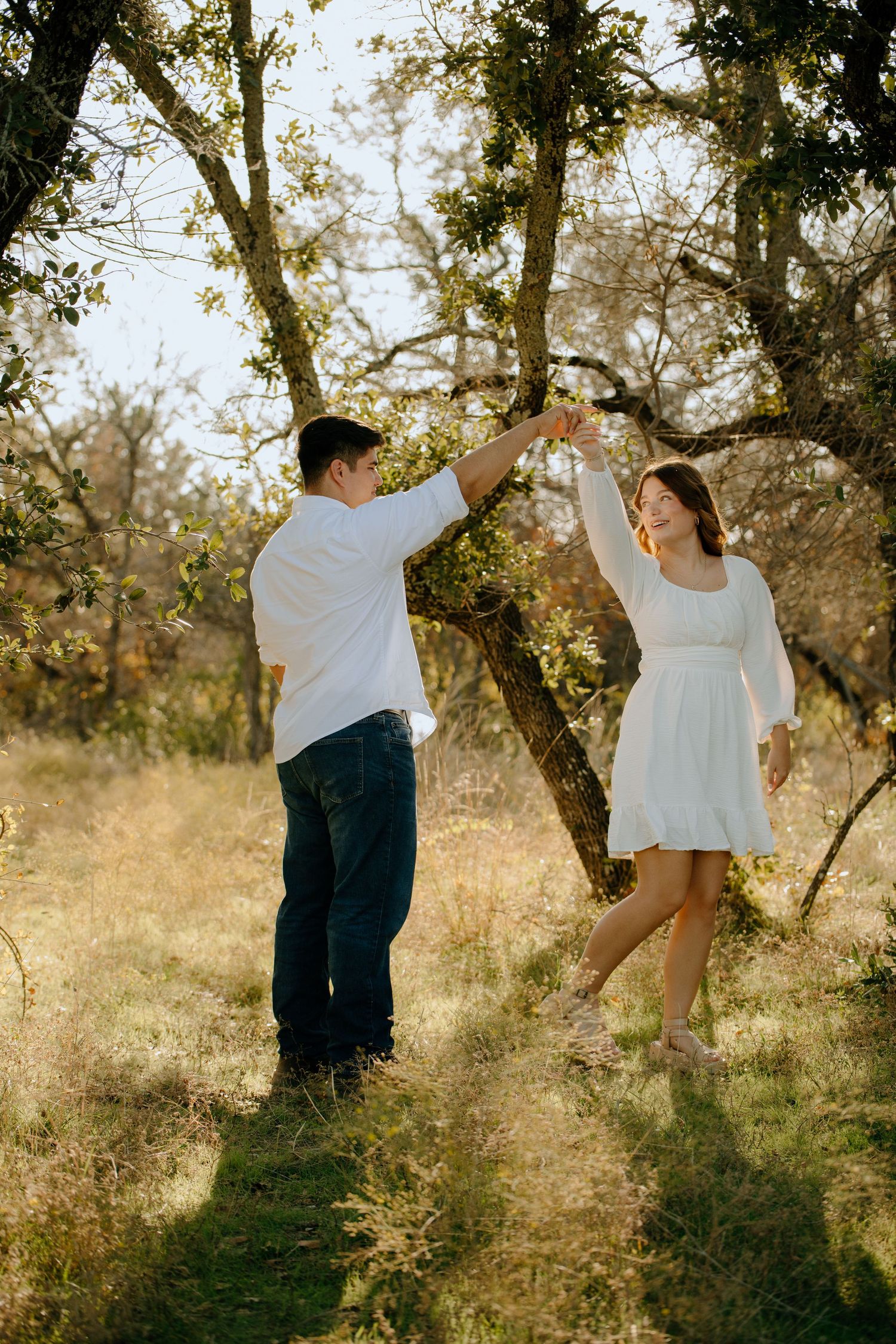 A romantic couple dances together in a sunlit meadow surrounded by trees during a golden hour photoshoot.