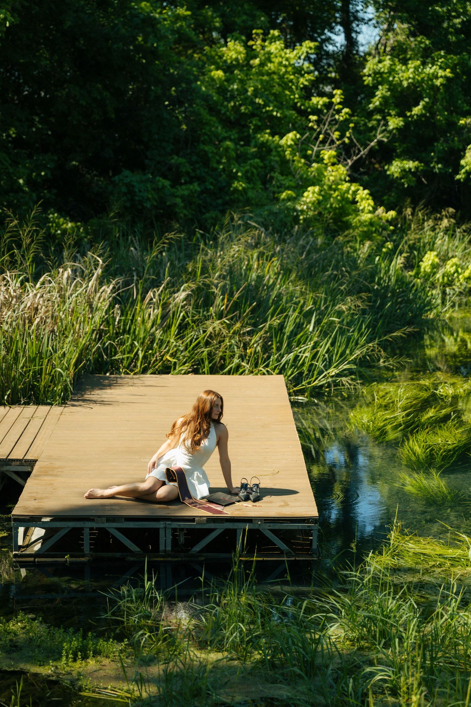 Woman in white dress sitting on wooden dock surrounded by lush green reeds and trees by a calm pond.