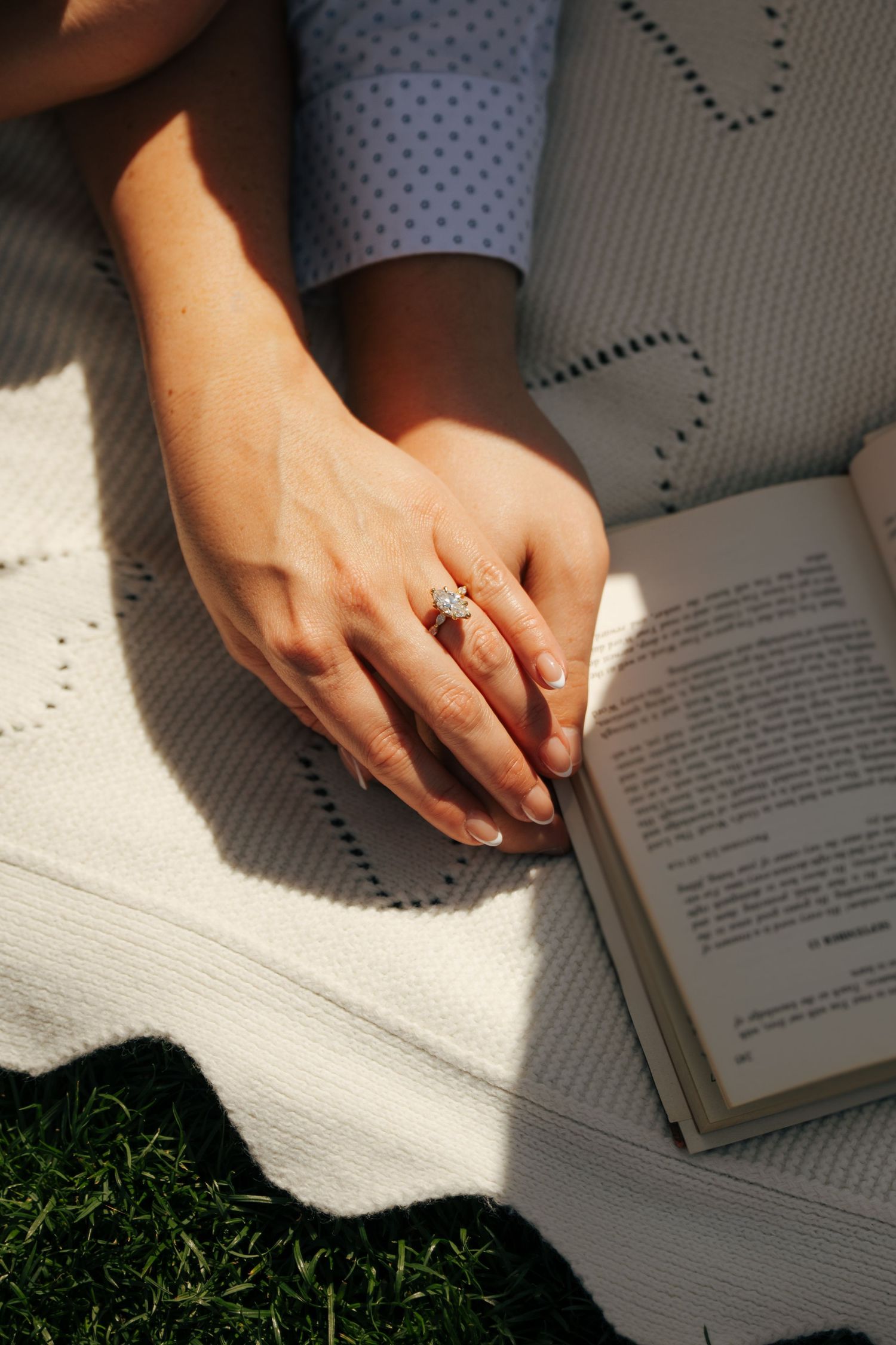 Close-up of hands with diamond engagement ring on white blanket beside open book on grass in sunlight.