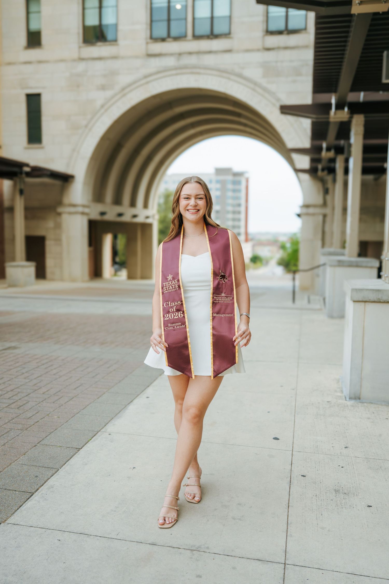 Smiling graduate in white dress and maroon stole walks confidently outside a stone archway building.