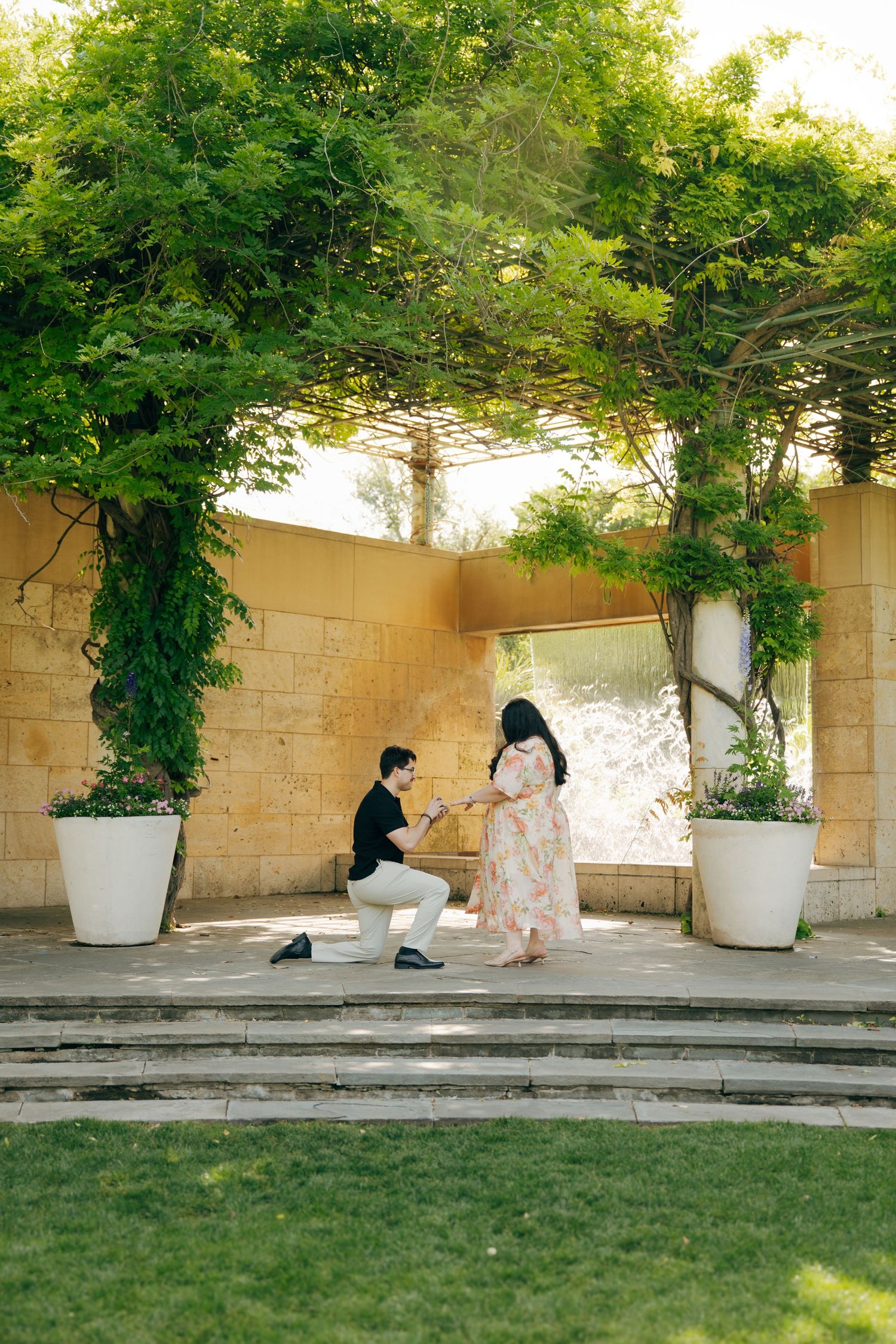 Man proposing on one knee to woman in floral dress under green pergola in elegant outdoor garden setting.