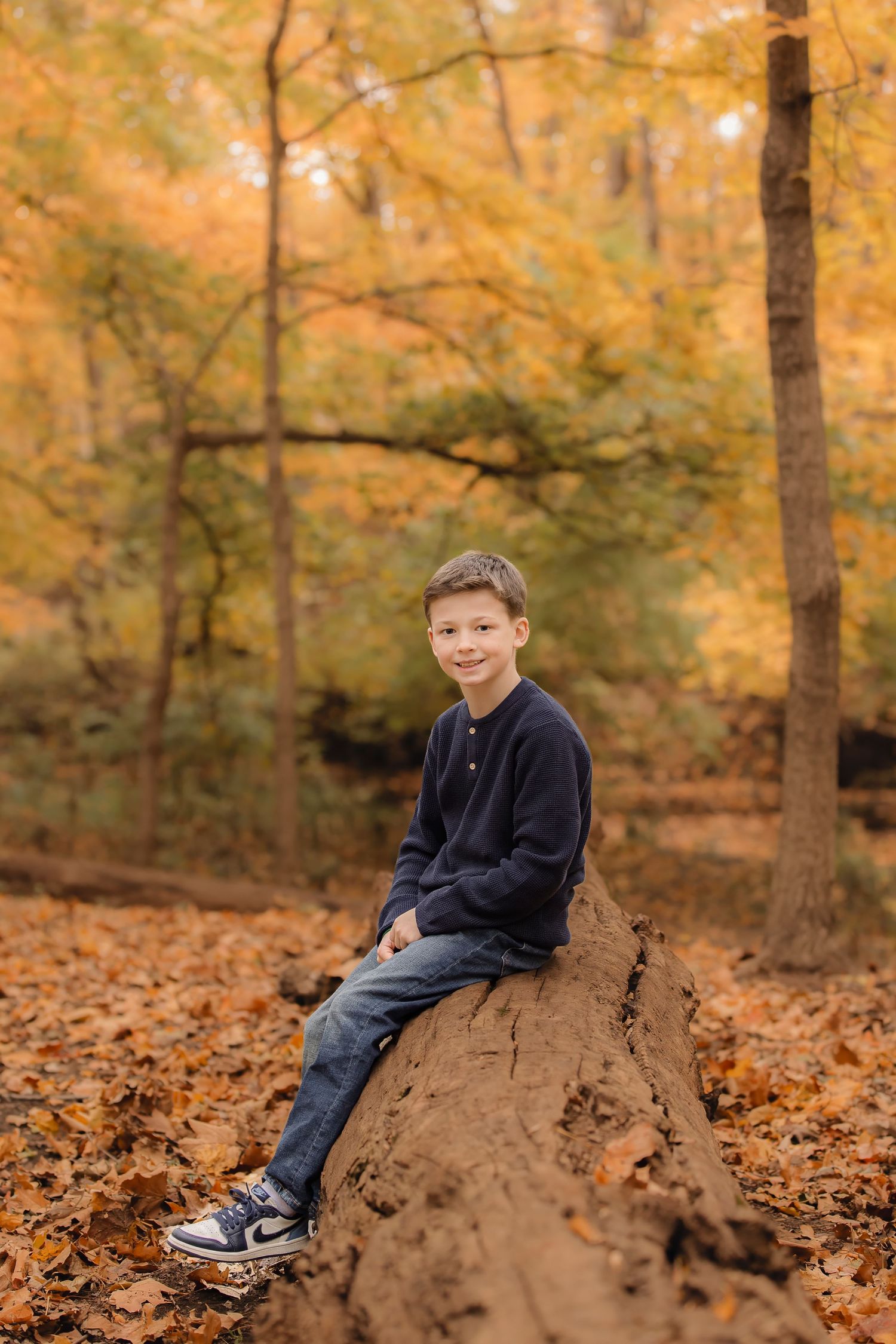 A child sits on a fallen log surrounded by golden autumn leaves in a forest setting.