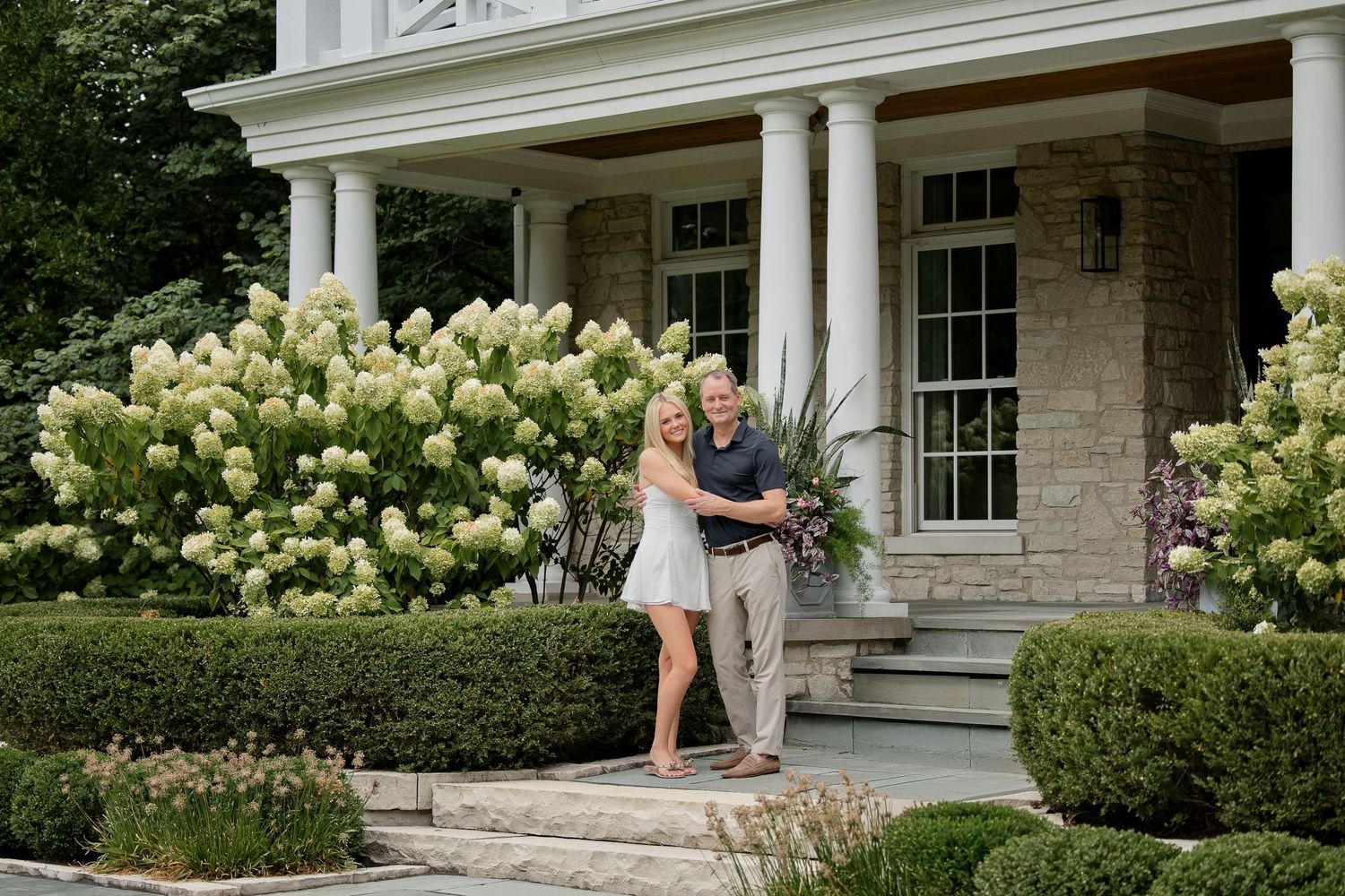 Couple smiling together in front of elegant stone home with white columns and blooming hydrangea bushes.