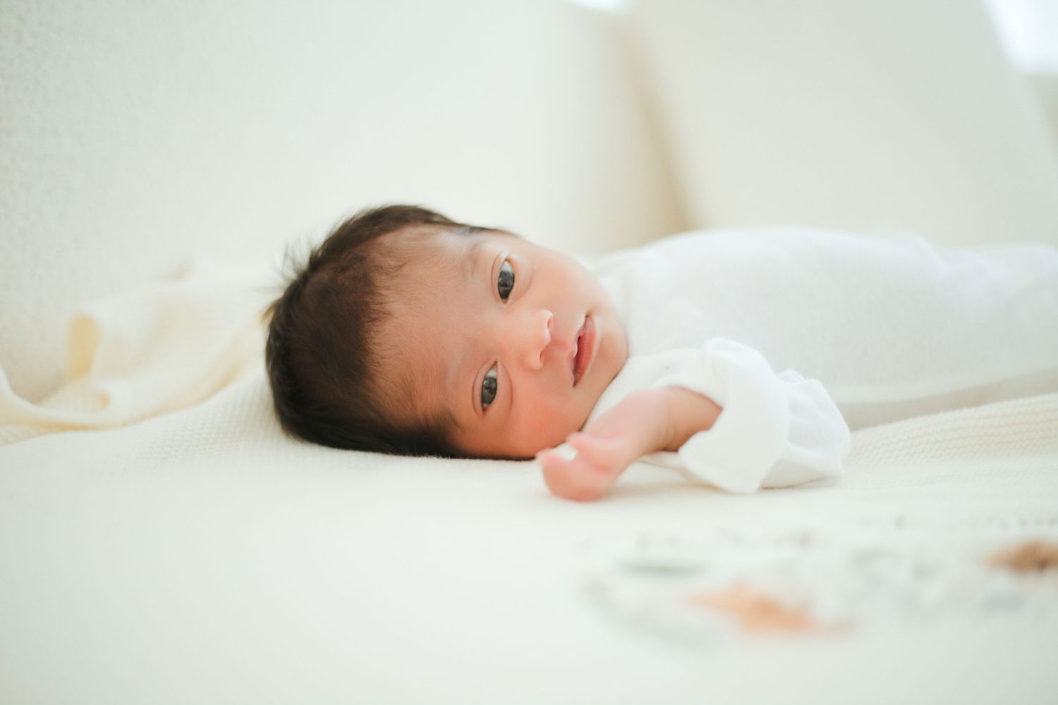 Newborn baby with dark hair lying on a white blanket, looking alert with tiny hands visible.