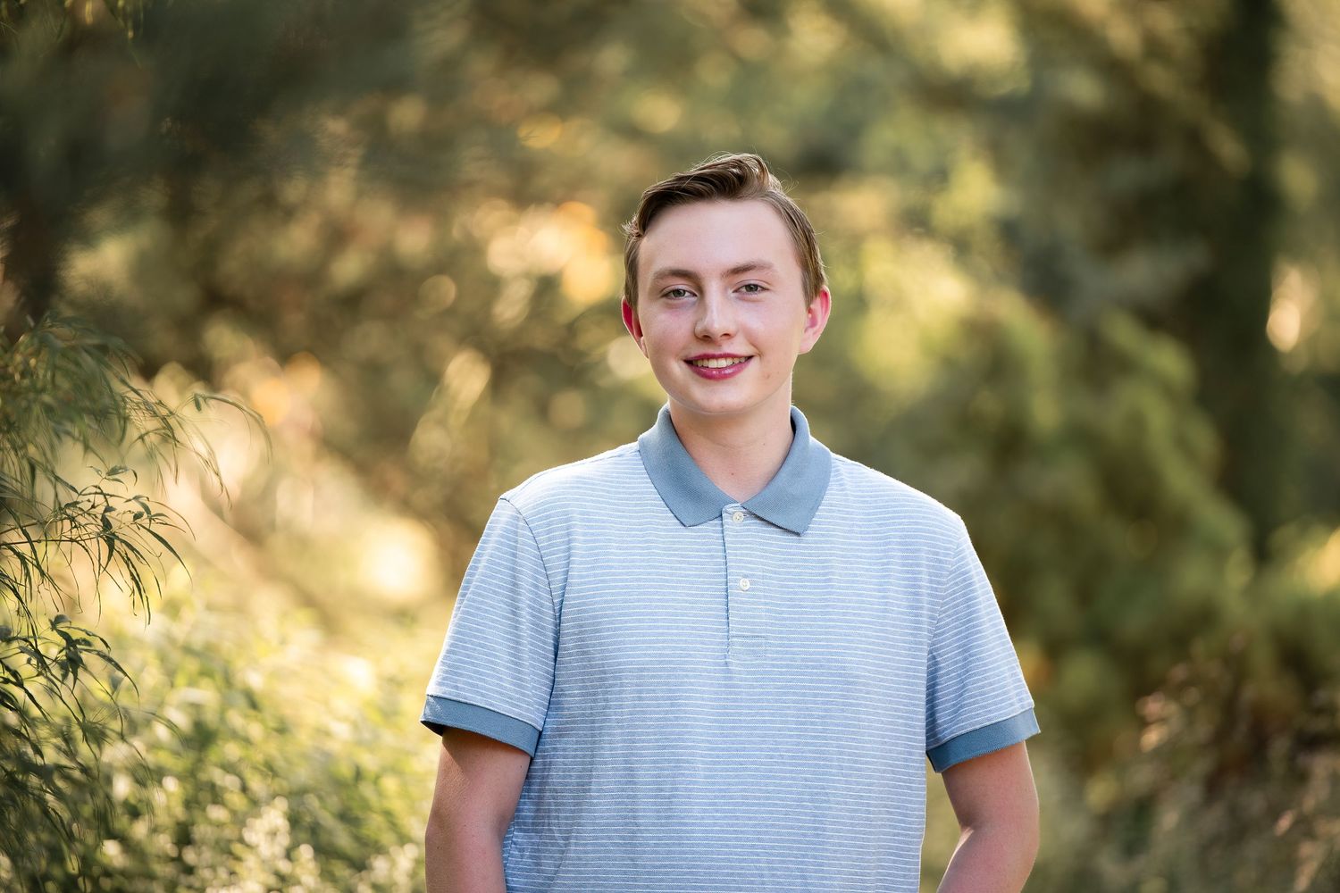 Smiling teenage boy in a light blue polo shirt posing outdoors among green trees in natural sunlight.