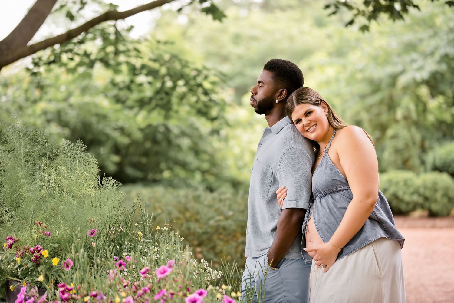 Couple posing in a lush garden with colorful flowers, woman smiling in gray dress, man in gray shirt.