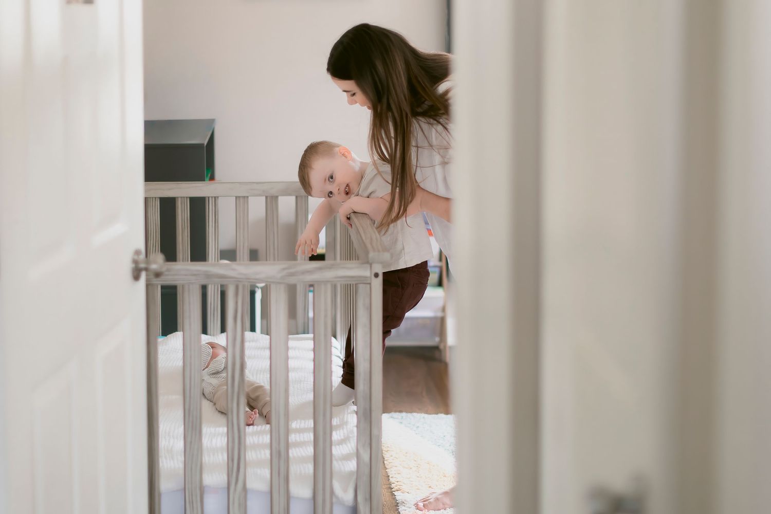 A mother tenderly interacts with her baby in a white crib inside a bright, minimalist nursery room.