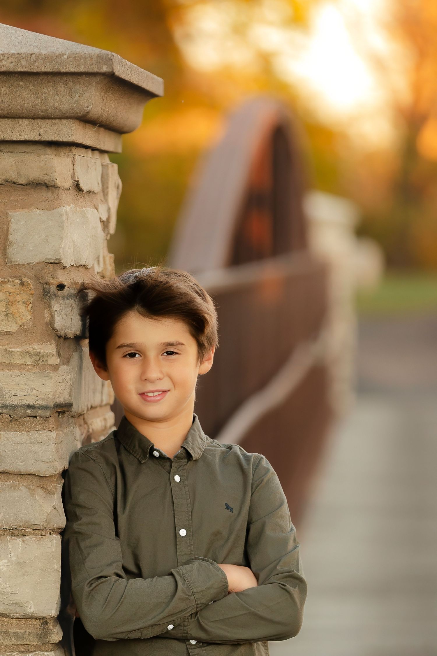 A child in an olive green button-down shirt poses against a stone pillar during golden hour.