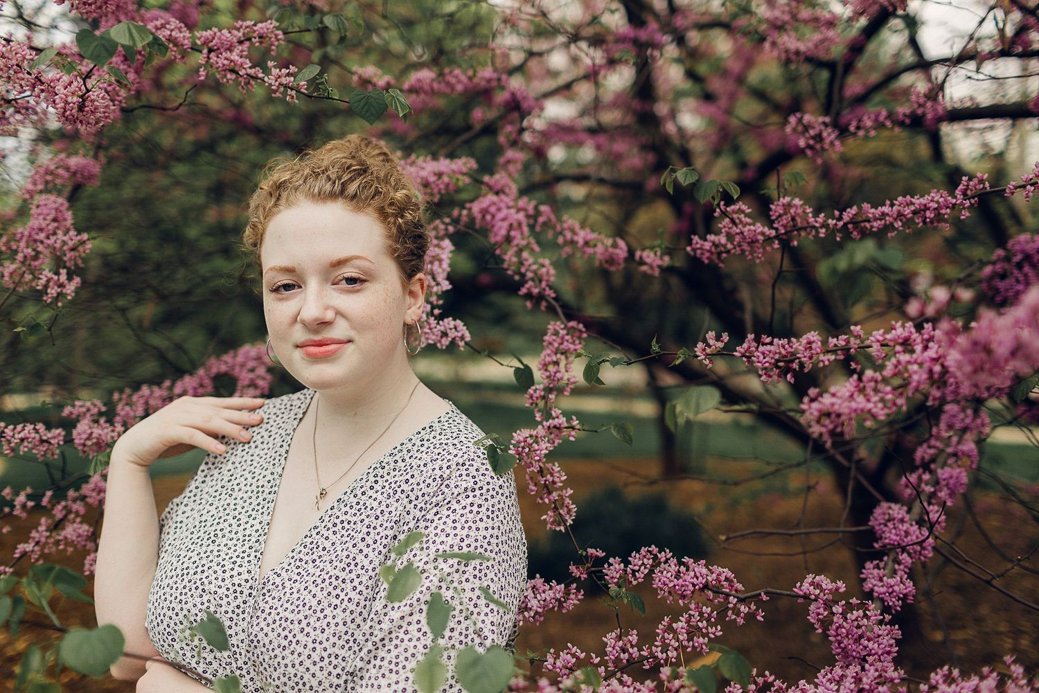 Woman in floral dress poses near blooming cherry blossom branches.