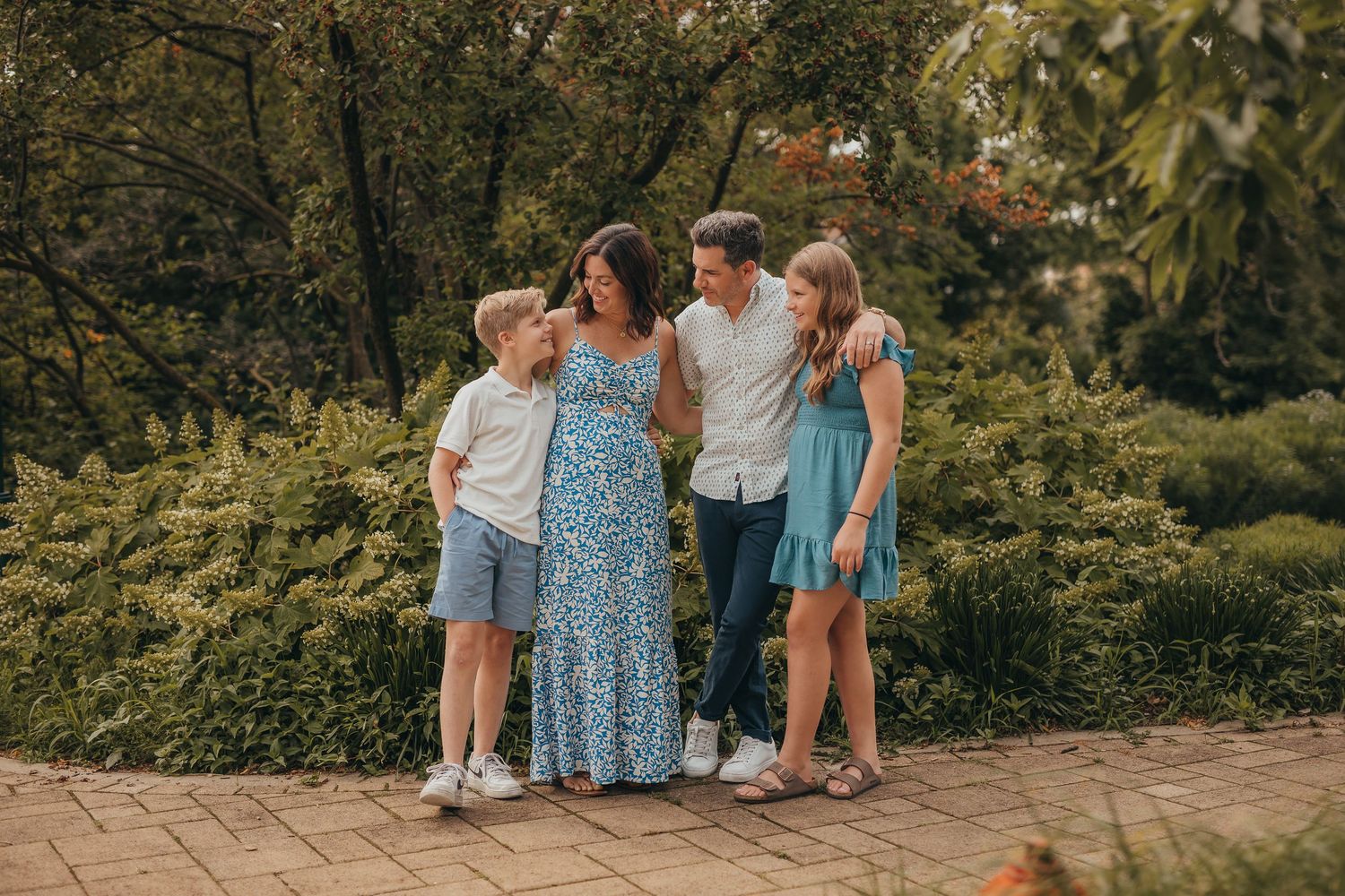Family of four posing outdoors in a lush garden, wearing casual summer outfits, smiling together on a brick path.