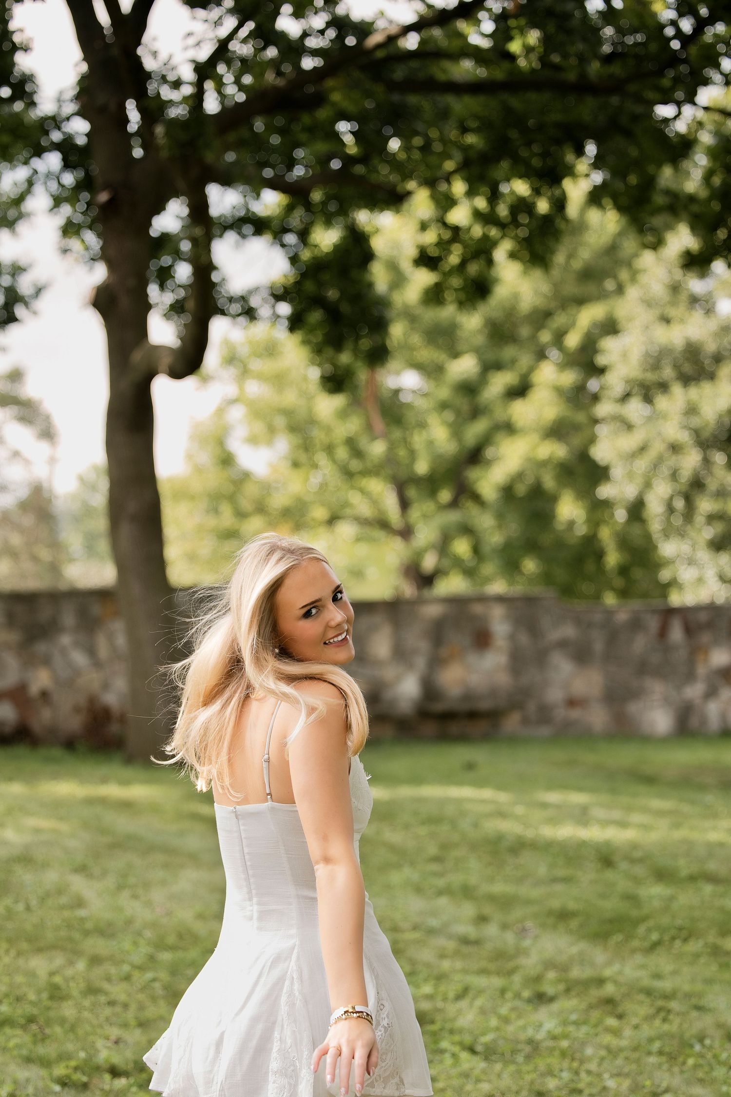 Smiling blonde woman in white sundress twirling in a lush green park with trees and stone wall background.