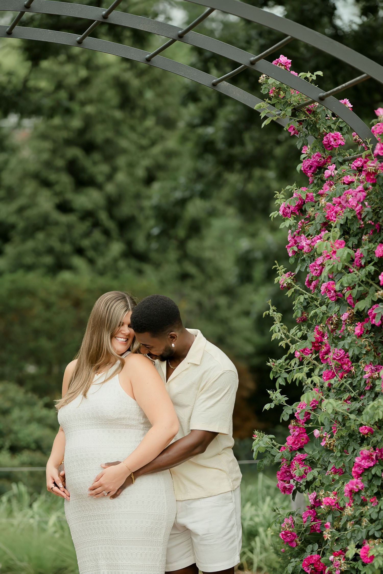 Pregnant couple in white outfits embracing under a floral garden arbor with pink climbing roses.