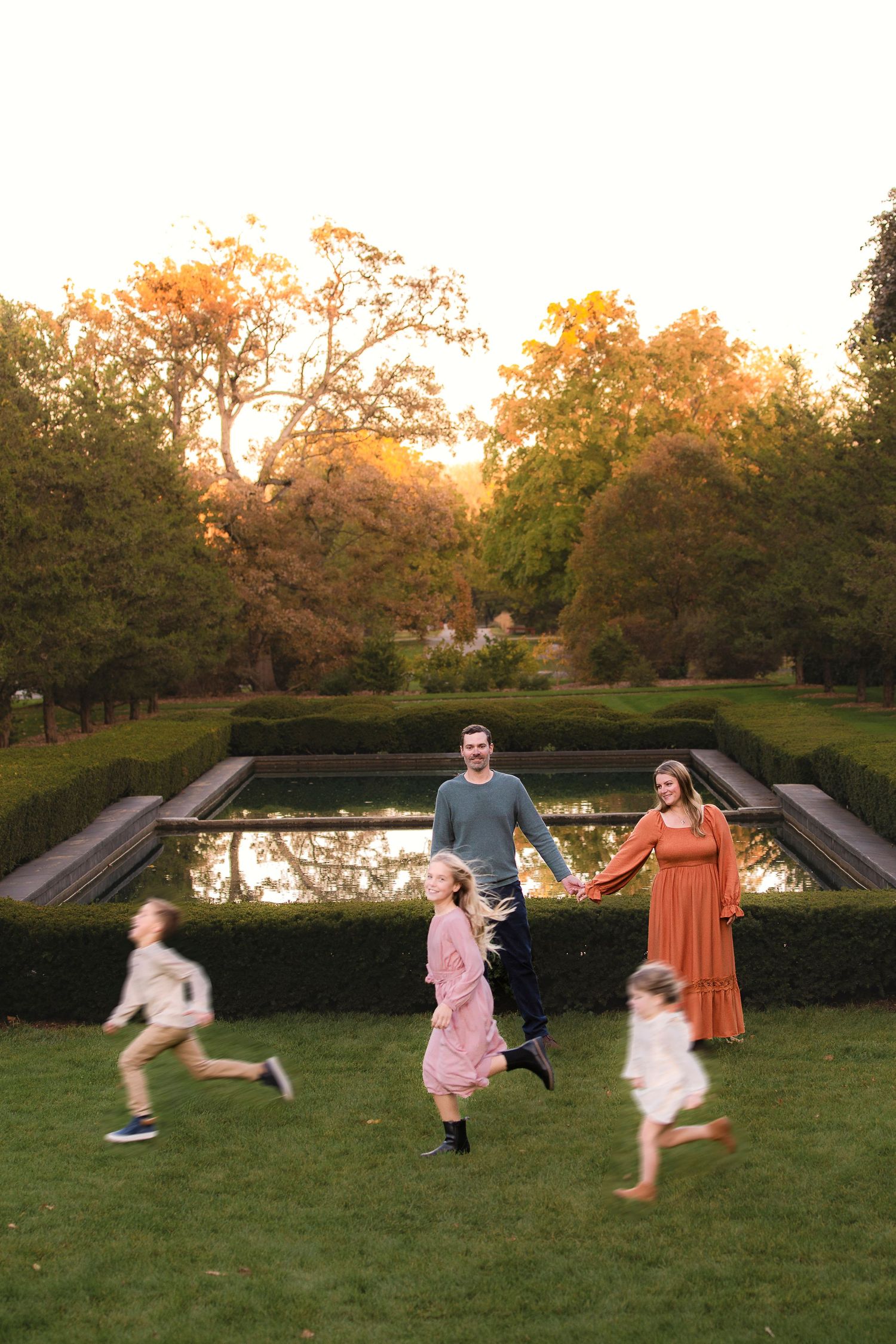 A group plays and runs together on a grassy lawn at sunset with trees in the background.
