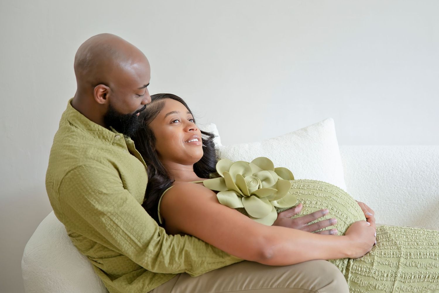 A loving couple in matching green outfits sit on a sofa, the man embracing his pregnant partner tenderly.