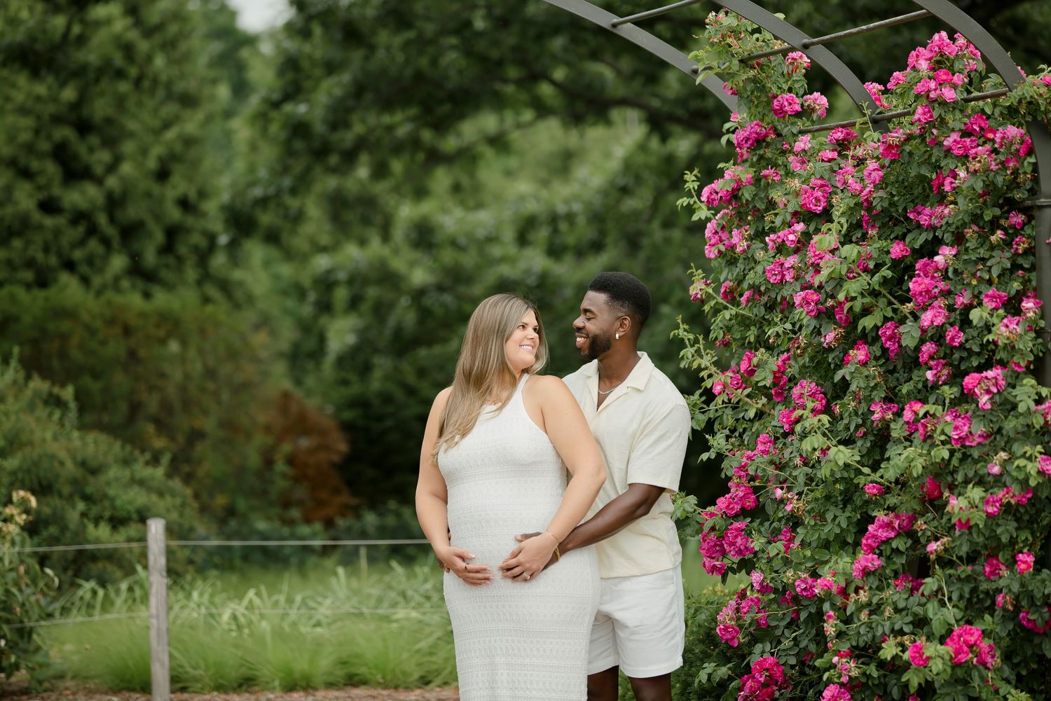 Interracial couple in white outfits pose by pink climbing roses, pregnant woman cradling baby bump in lush garden.