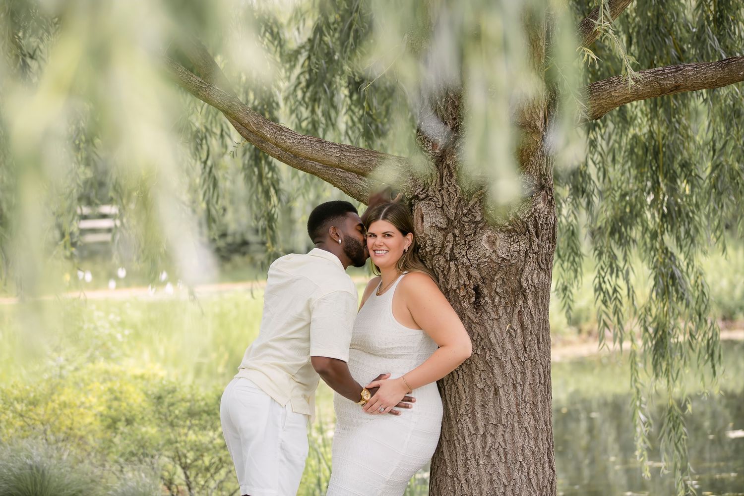 Couple in white wedding attire sharing a romantic moment under a weeping willow tree by a pond.