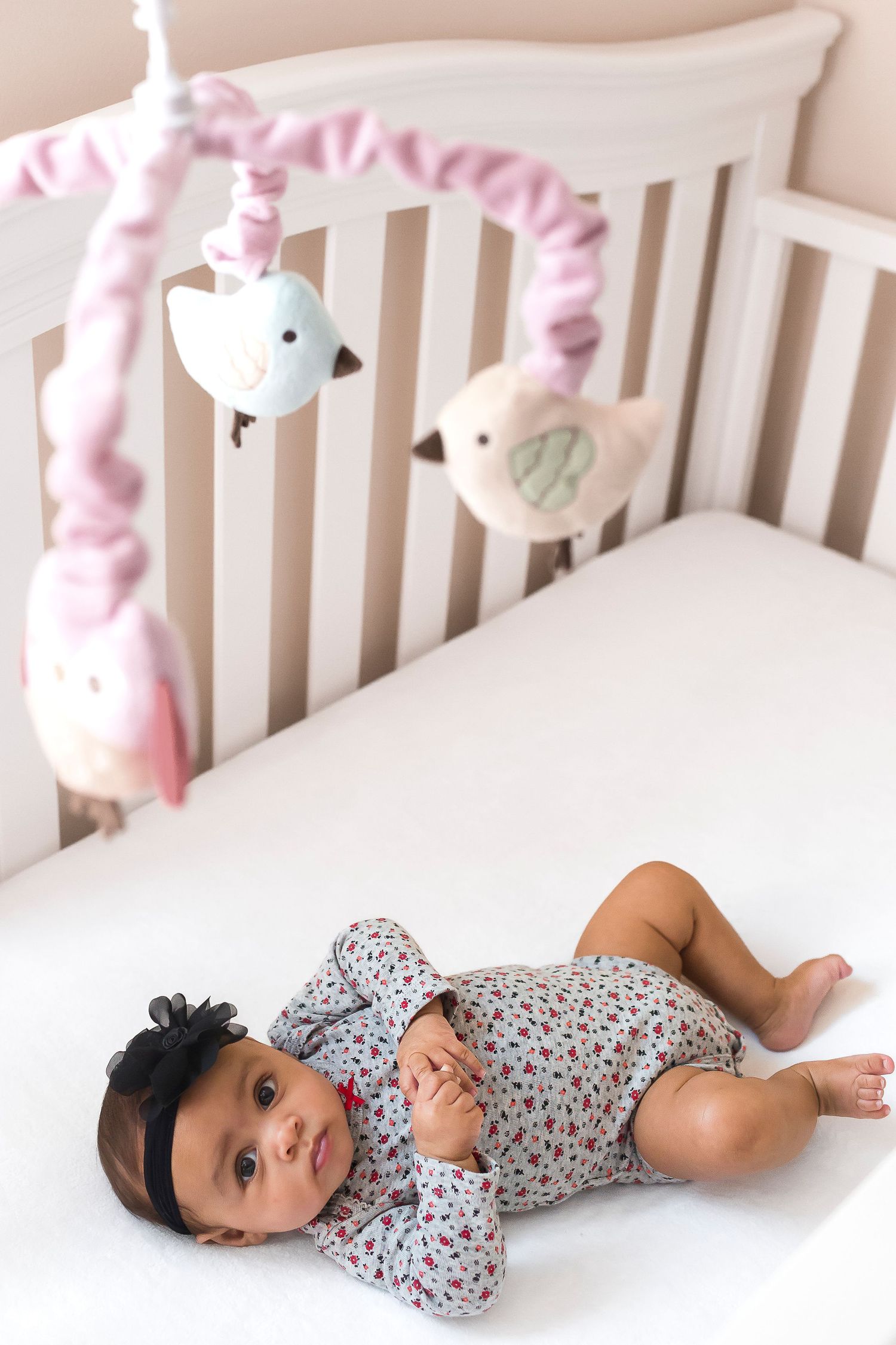 A baby in a floral onesie lies peacefully in a white crib surrounded by plush toys.