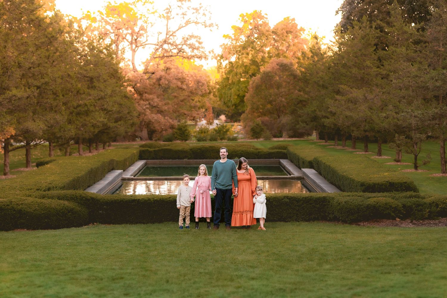 A group stands at sunset overlooking a reflecting pool surrounded by manicured hedges and autumn trees in a garden setting.