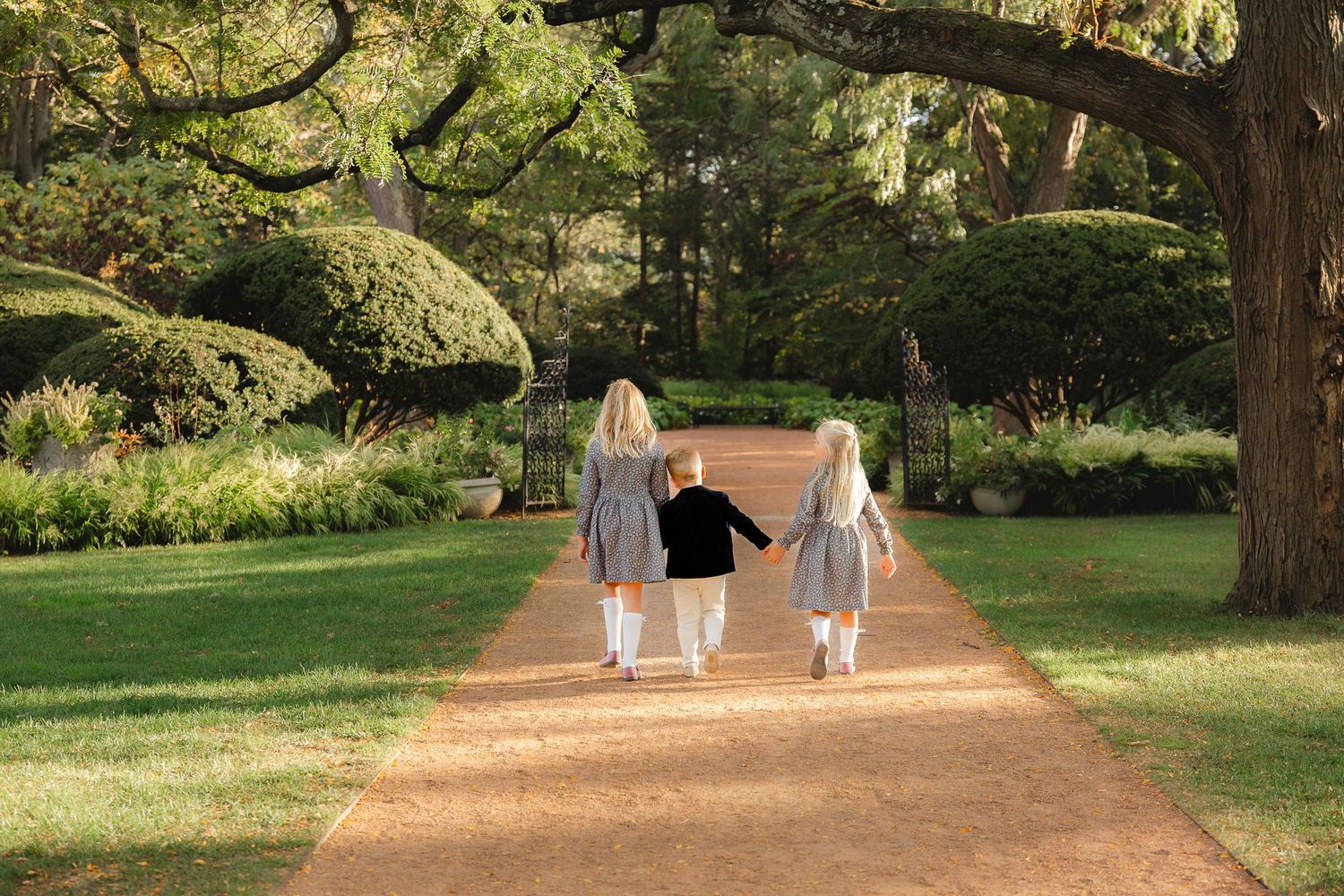Three people in white outfits walk along a sunlit garden path surrounded by lush greenery and tall trees.