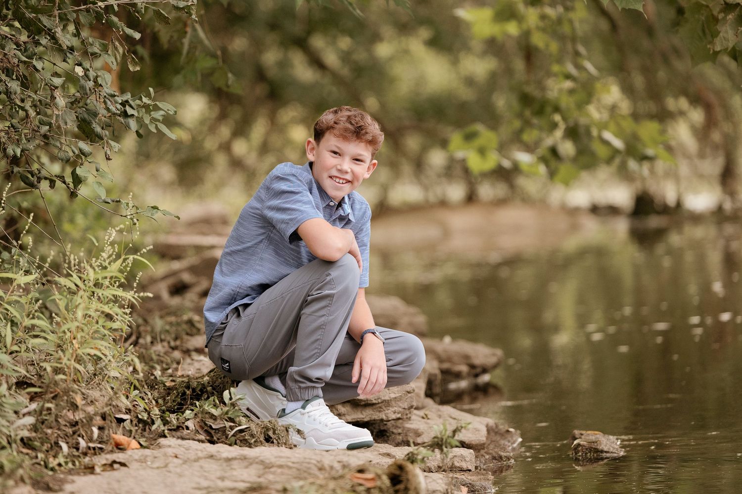 A smiling boy sits on rocks by a peaceful creek, surrounded by lush green trees, wearing a blue shirt and gray pants.