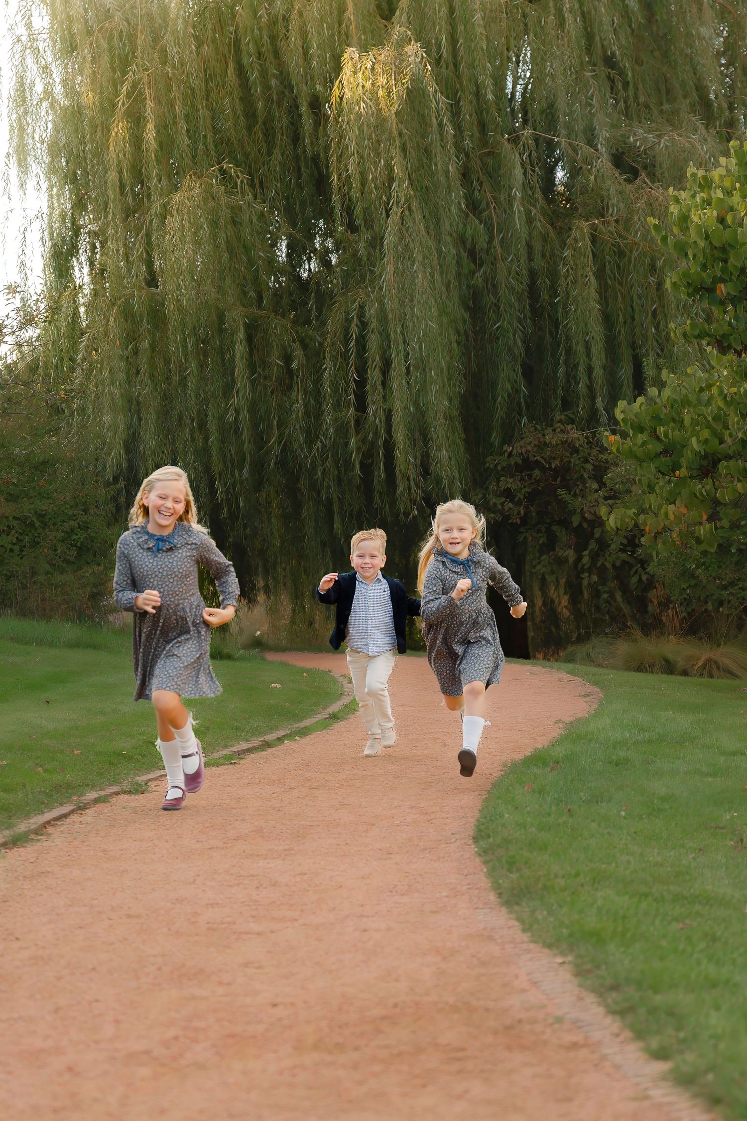 Children run playfully down a dirt path lined with drooping willow trees in a park setting.