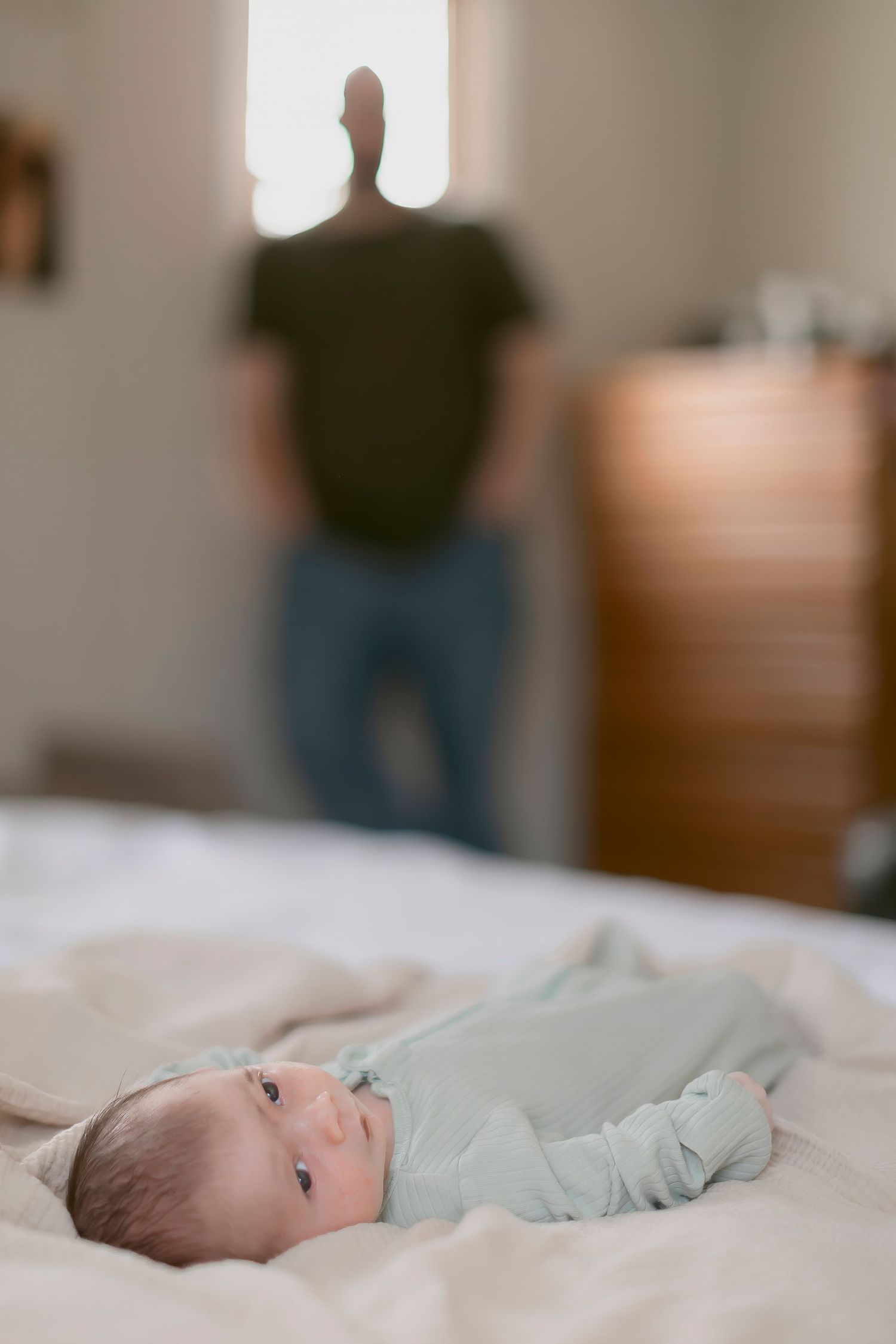 Newborn baby in mint onesie lying on white bed with blurred parent standing by window in background.