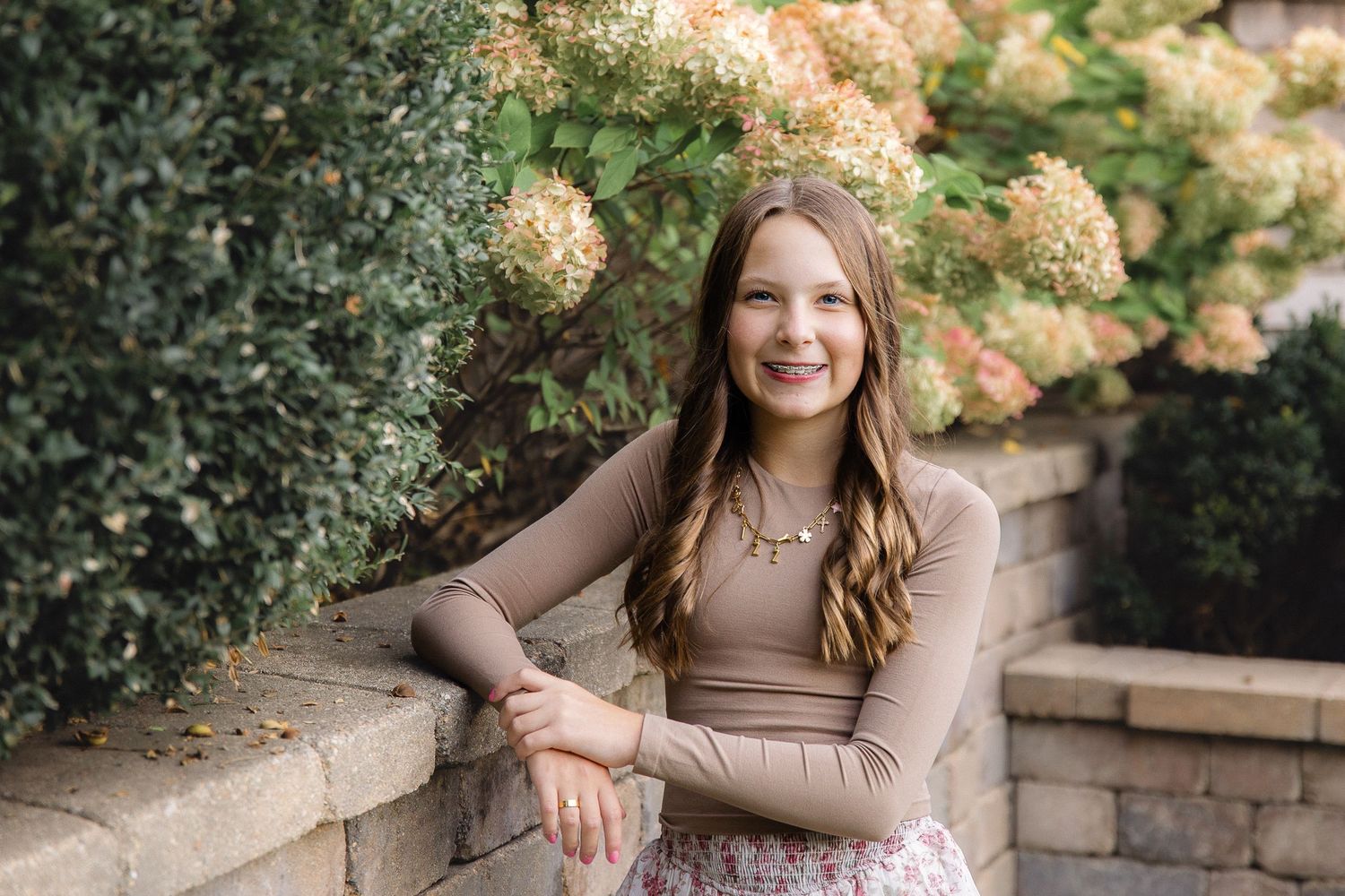 A portrait of someone in a brown sweater posed against cream hydrangeas and stone garden walls.