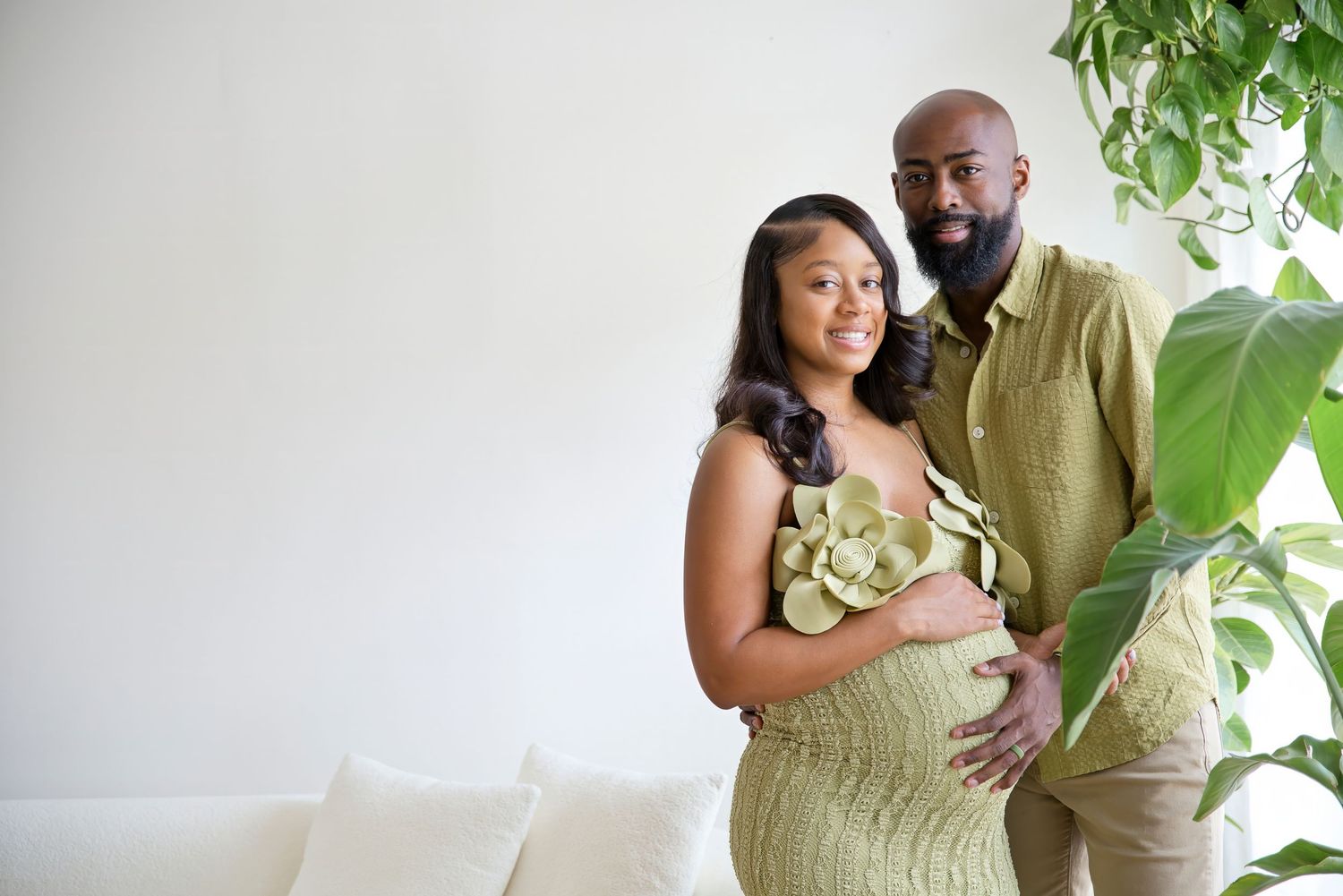 A couple poses for a maternity photo, the pregnant woman in a green floral dress, her partner beside her in an olive shirt.