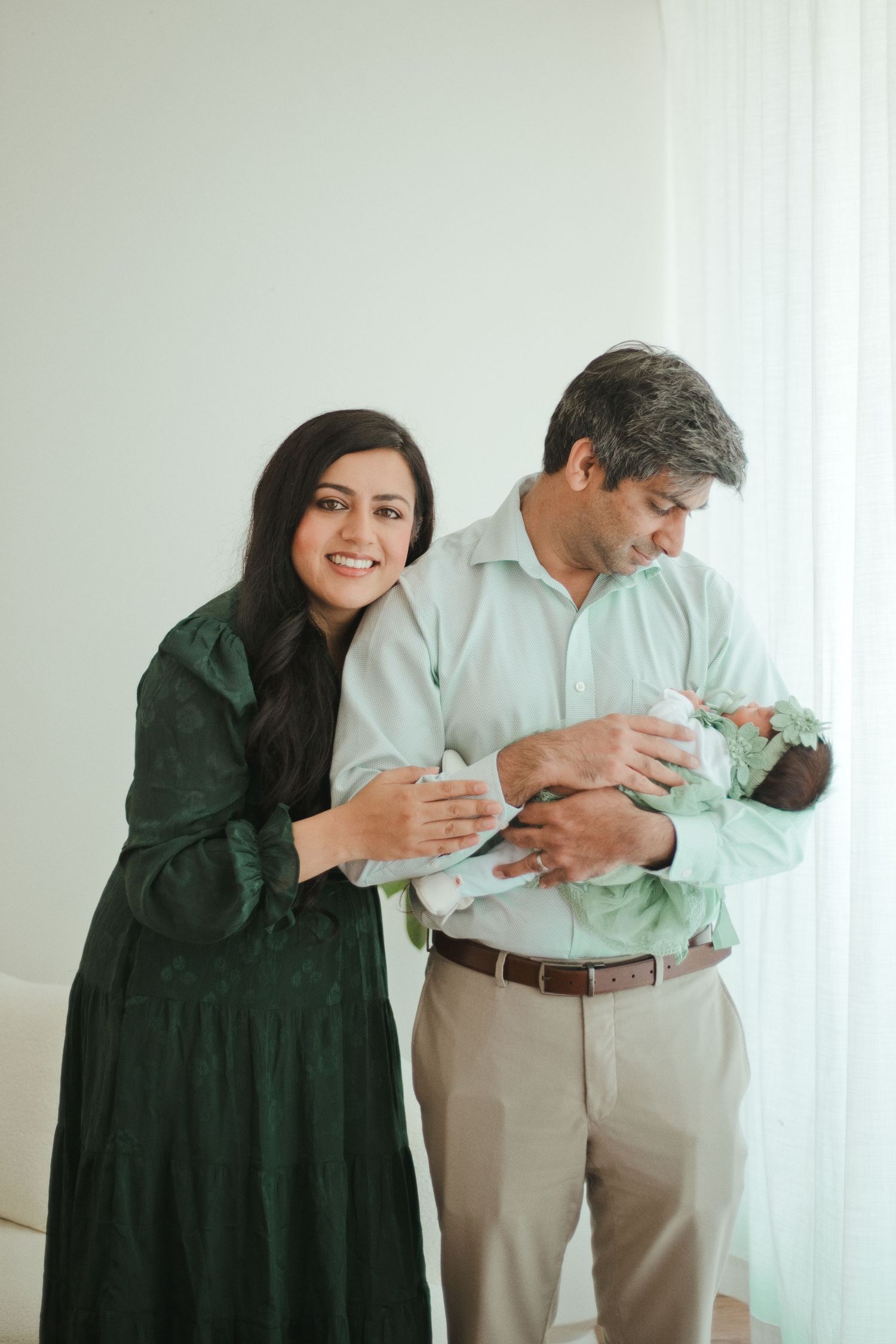 A smiling couple in a bright room holds their newborn baby, the woman in green dress, man in mint shirt.